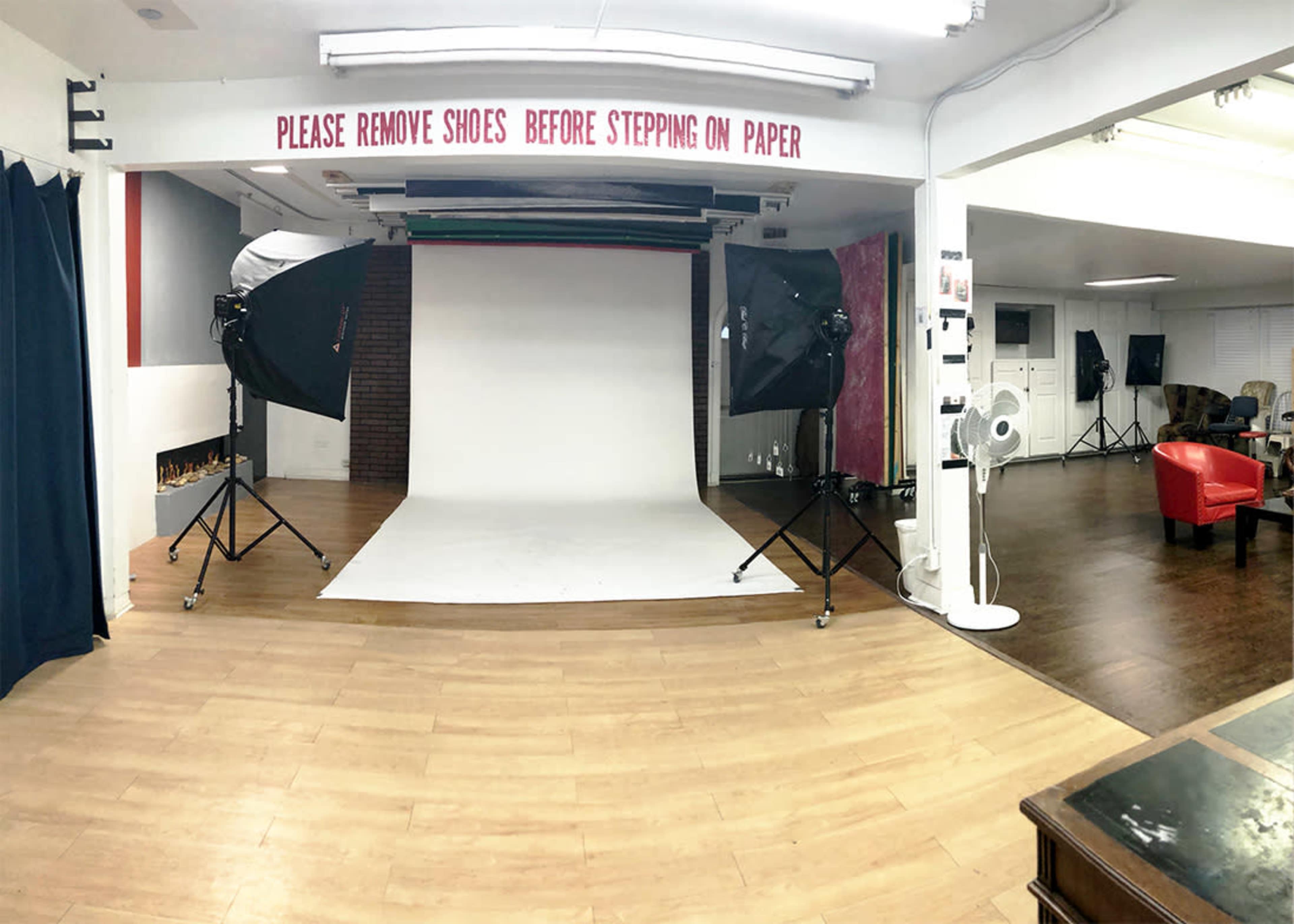 The image shows a photography studio with a clean, white backdrop surrounded by studio lights and a sign instructing visitors to remove their shoes before stepping on the paper.