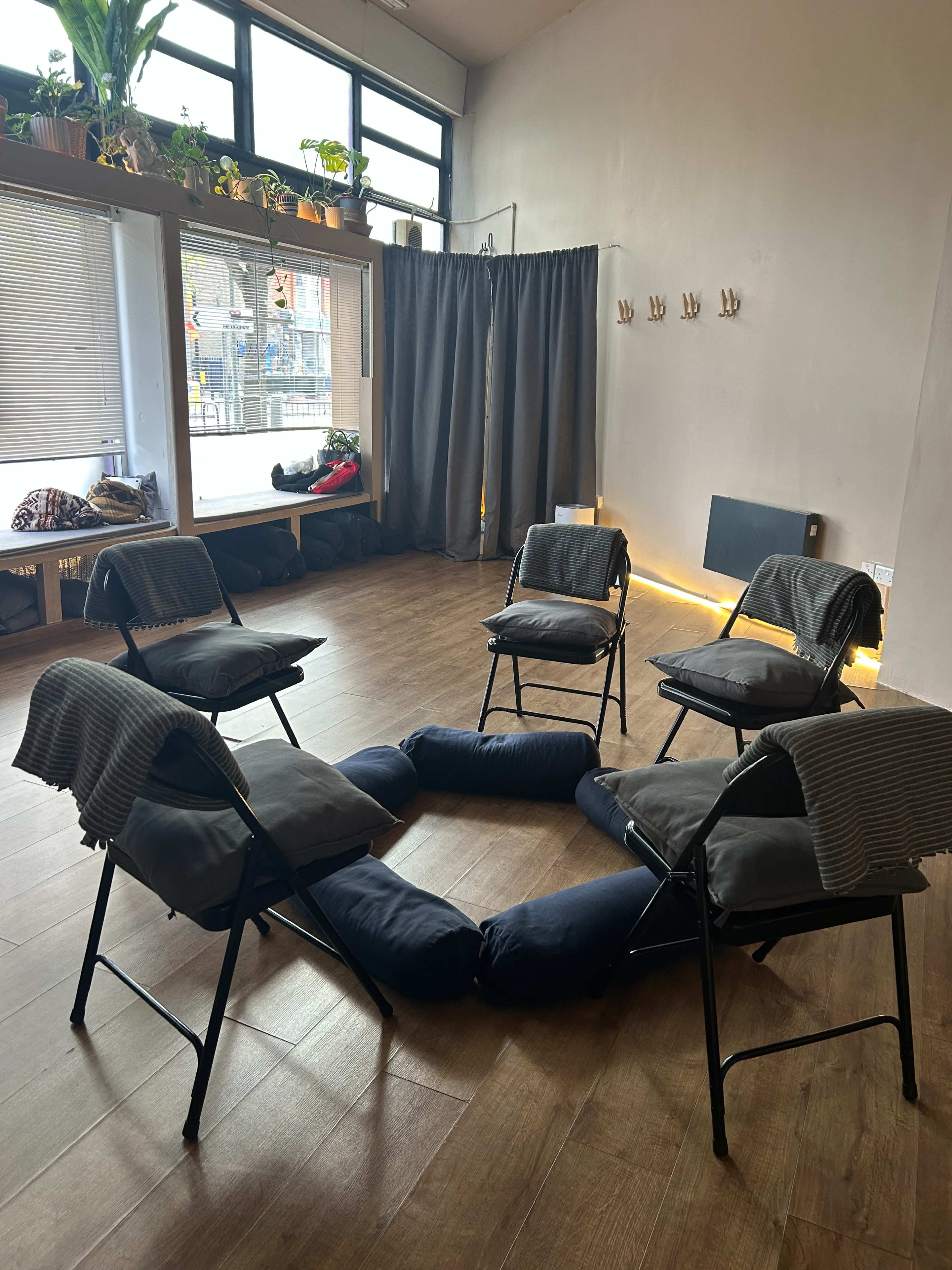 A circle of folding chairs with cushions is arranged on a wooden floor, surrounded by a cozy, well-lit space featuring plants and curtains.