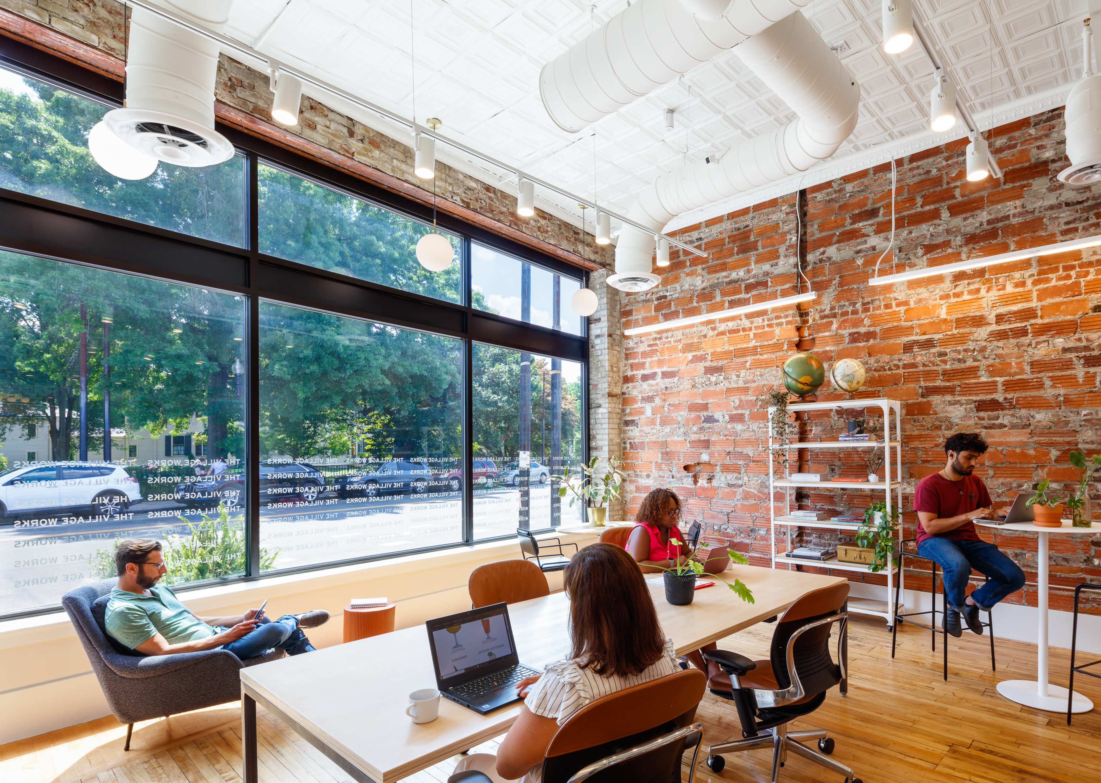 A modern office space features a mix of individuals working at a long table, chairs, and standing desks, surrounded by large windows and exposed brick walls.