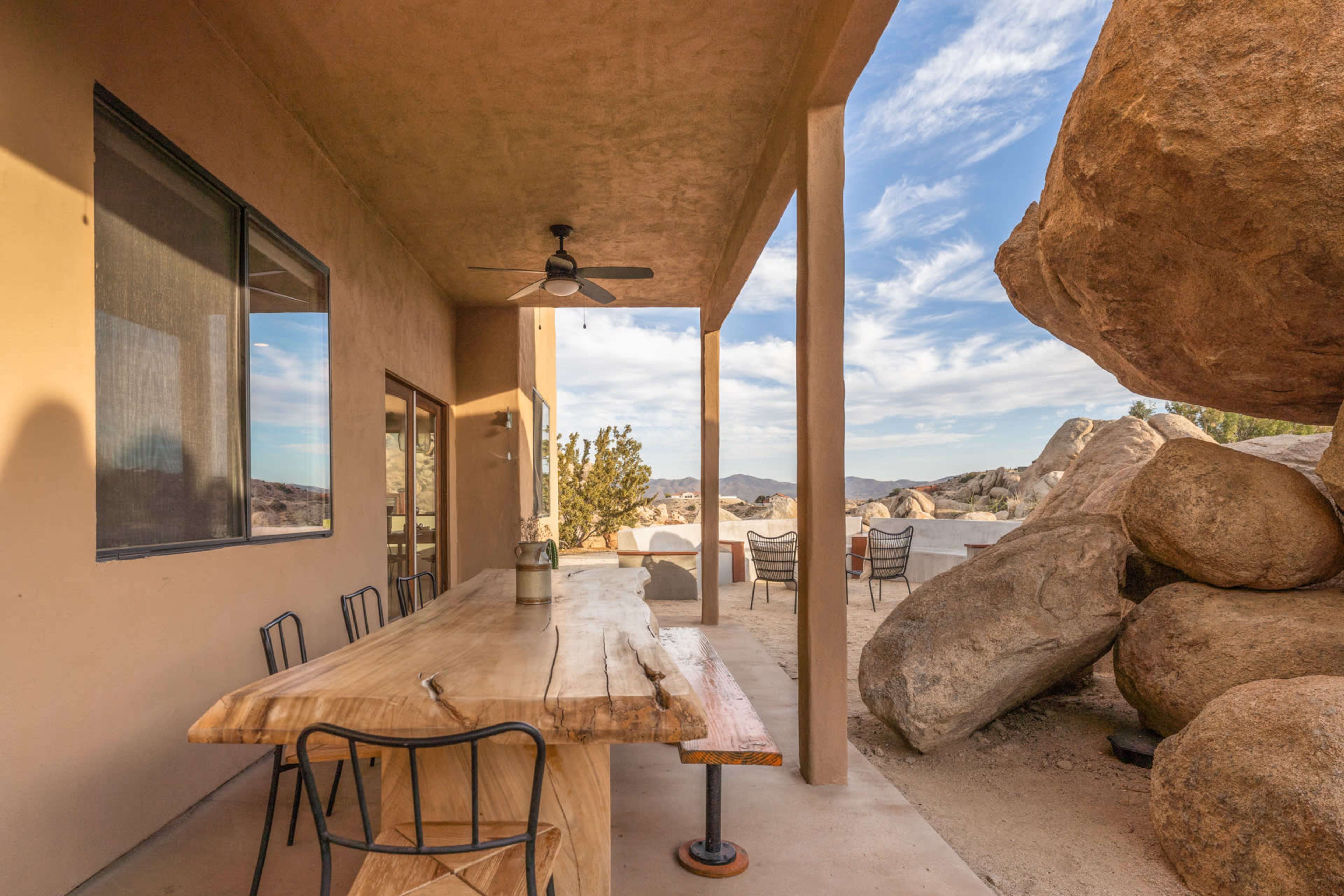 A patio with a large wooden table and chairs, adjacent to a rock formation and desert landscape.