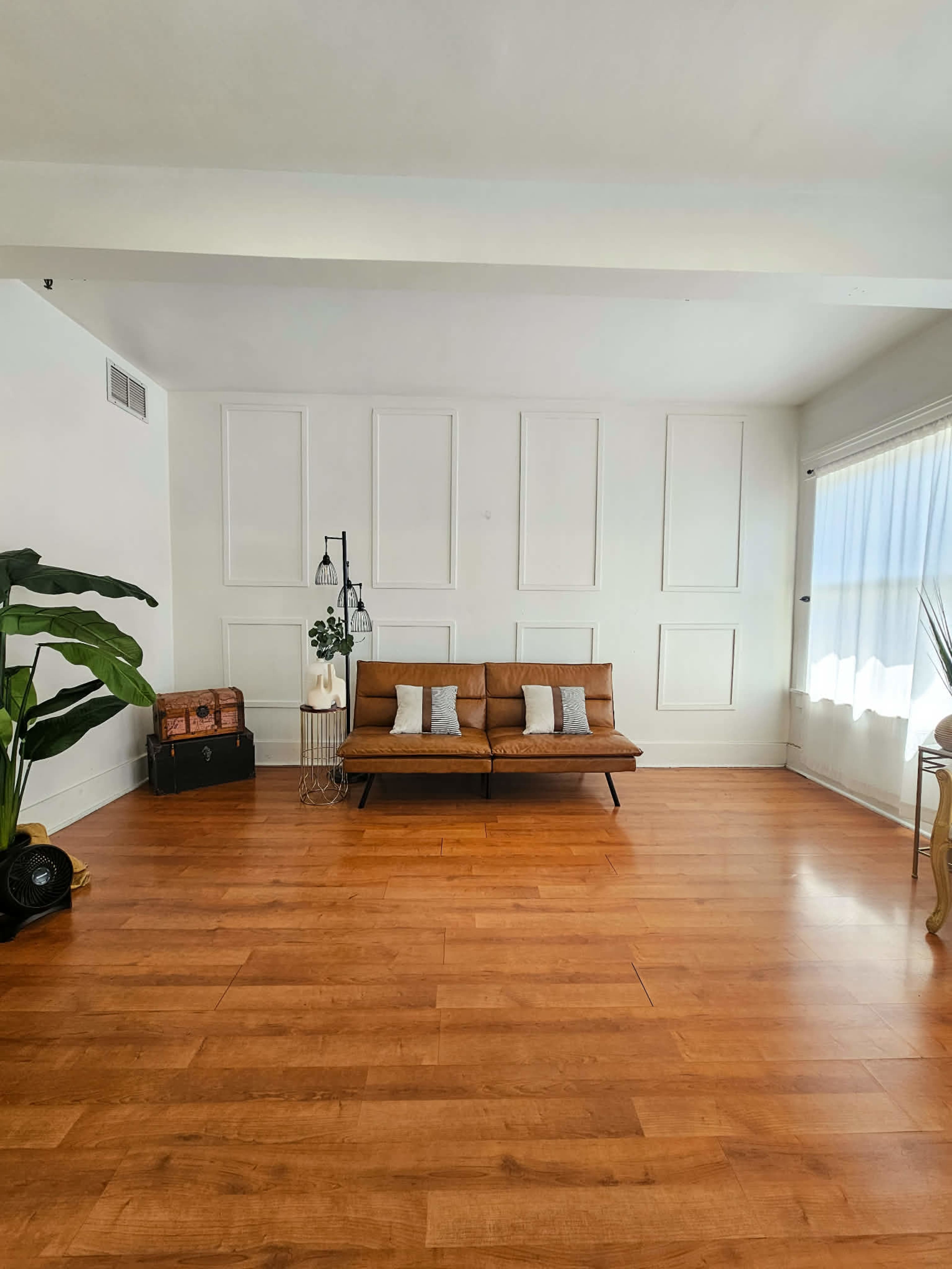 The image shows a minimalist living room with a brown sofa, a large indoor plant, a standing lamp, and wooden flooring.