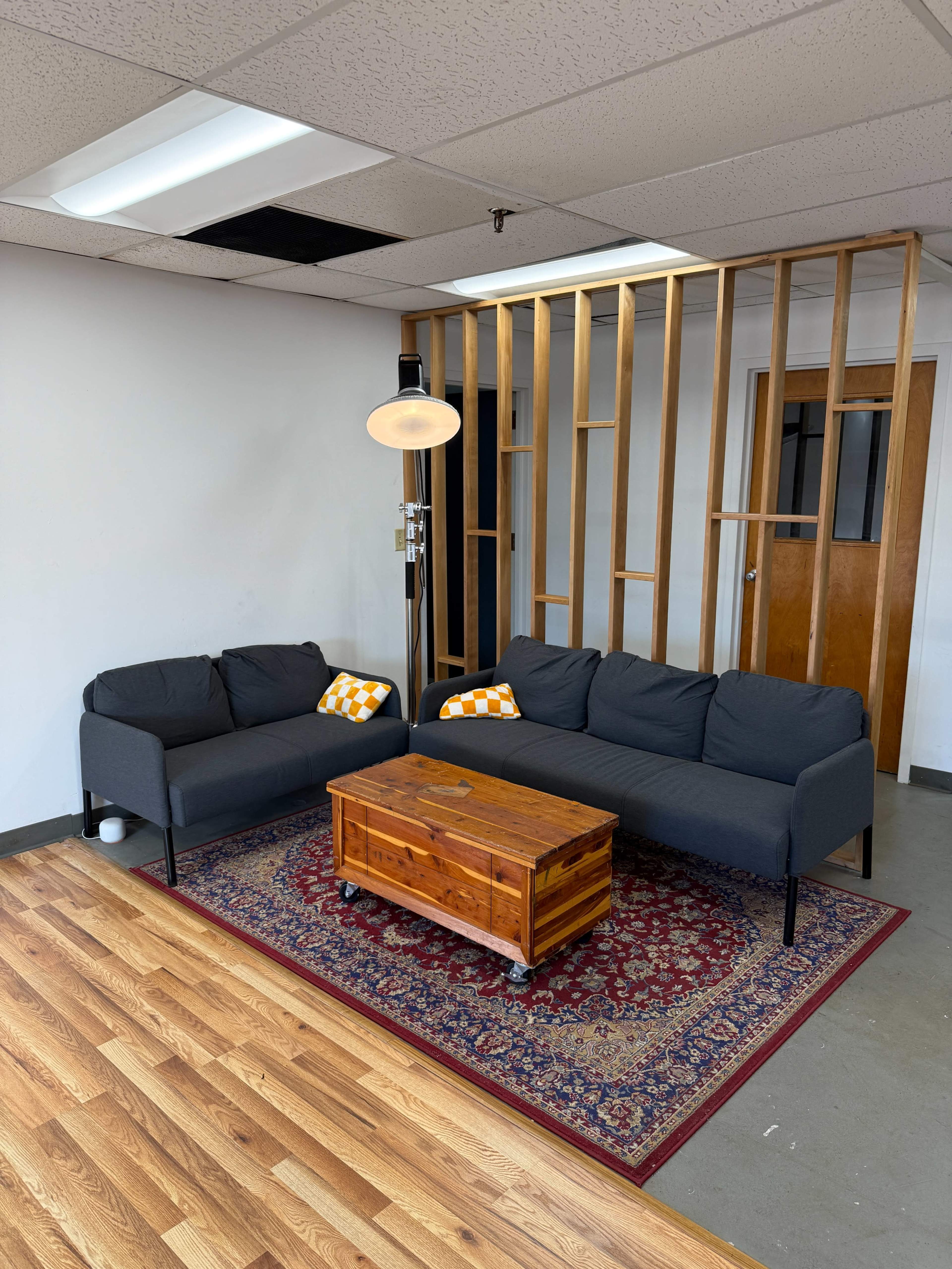 The image shows a minimalist office space featuring a dark gray sectional sofa with orange and white accent pillows, a wooden coffee table on wheels, and a patterned area rug.