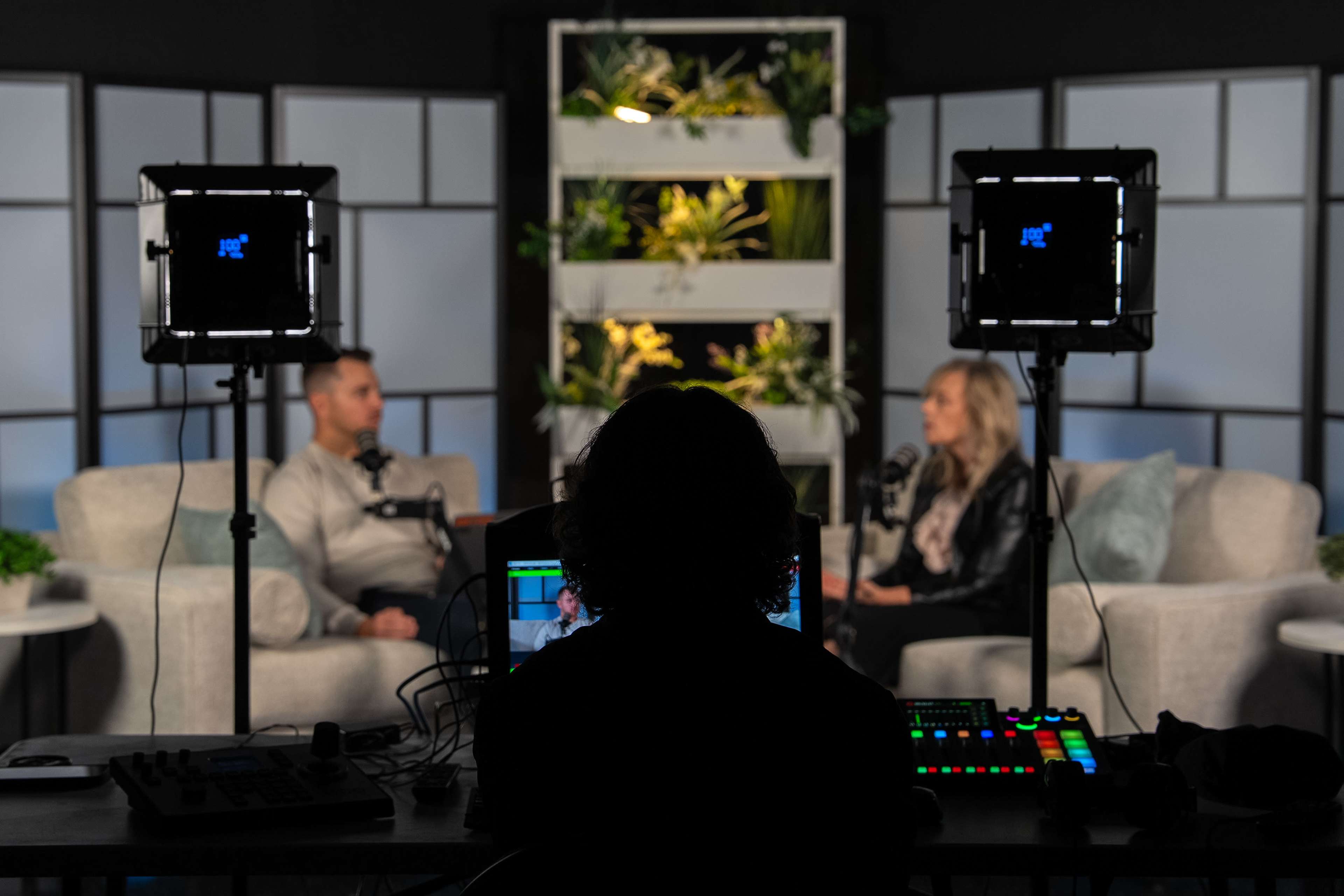 A person sits at a control panel in front of two individuals engaged in conversation on a set designed with greenery in the background.