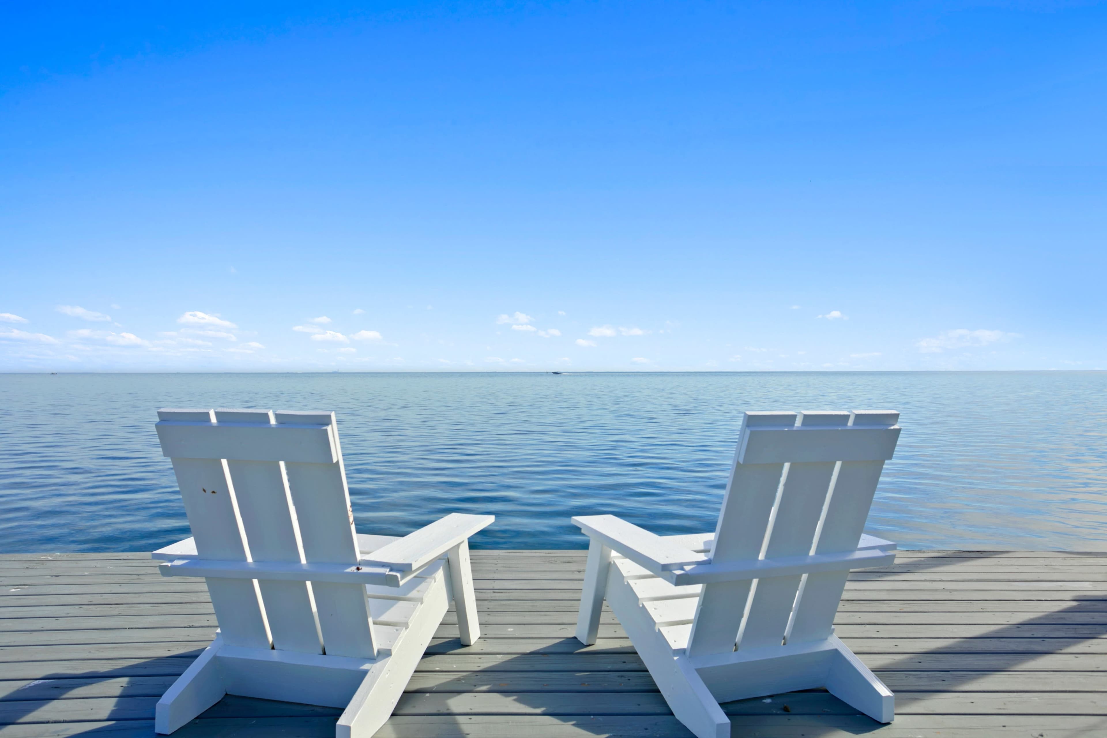 Two white Adirondack chairs are positioned on a wooden dock overlooking a calm body of water under a clear blue sky.