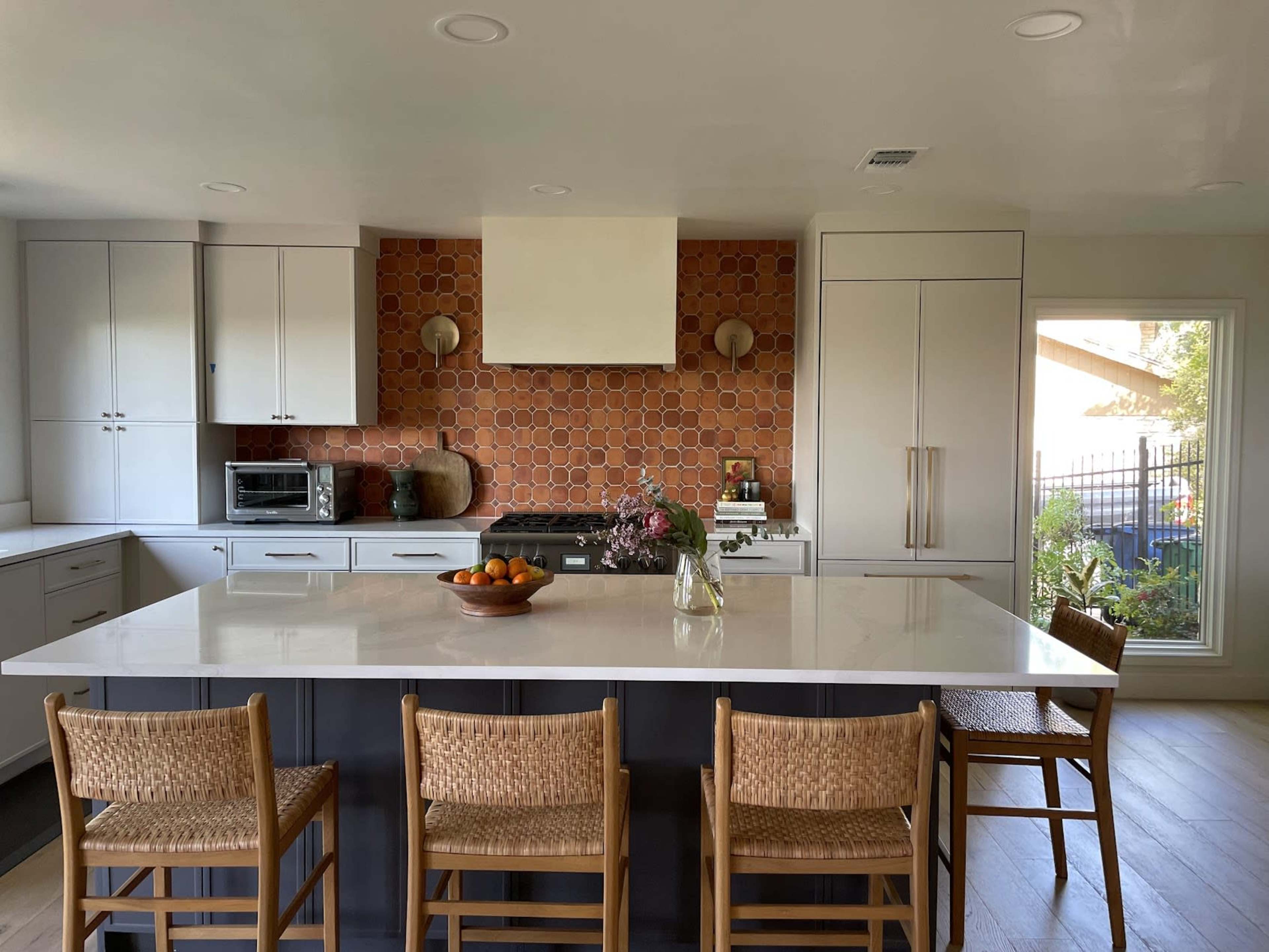 A modern kitchen features a large island with wooden stools, a hexagonal tile backsplash, and a view of an outdoor space through a window.