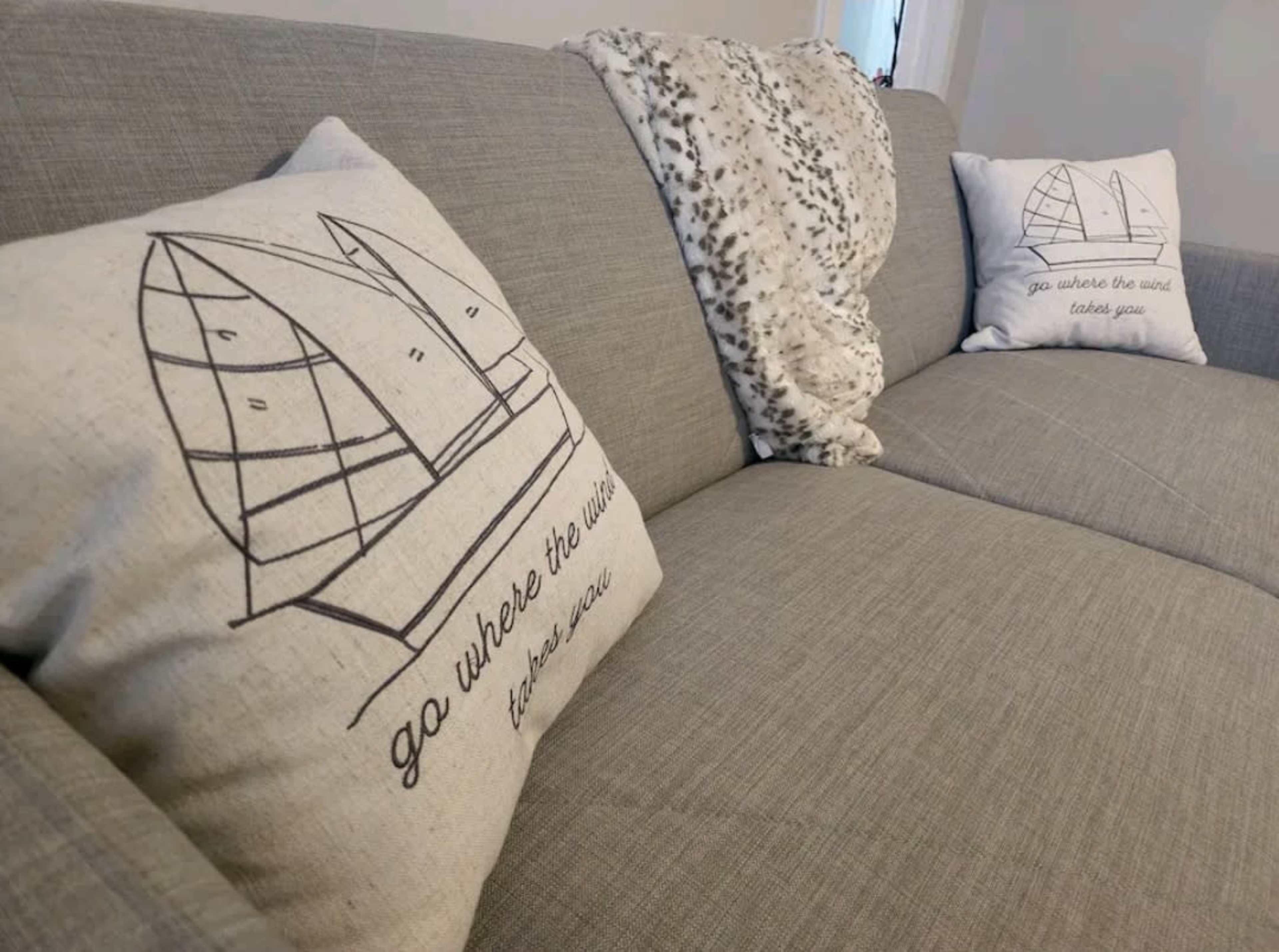 The image shows a gray sofa with two decorative pillows featuring an illustration of a sailboat and a phrase printed on them, along with a cozy throw blanket draped over the armrest.