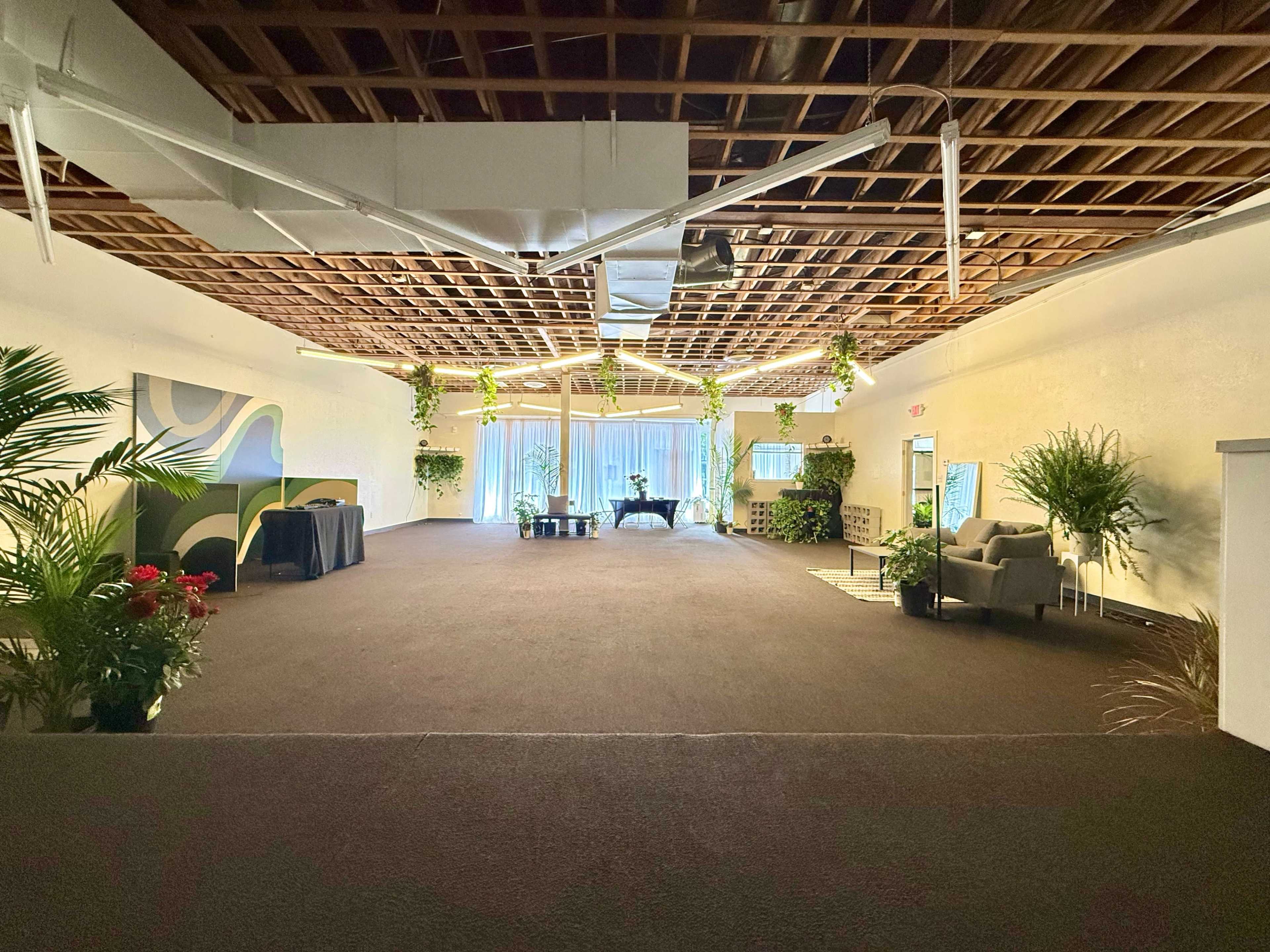 The image shows a spacious interior with a high ceiling, featuring plants, furniture, and natural light coming through large windows.