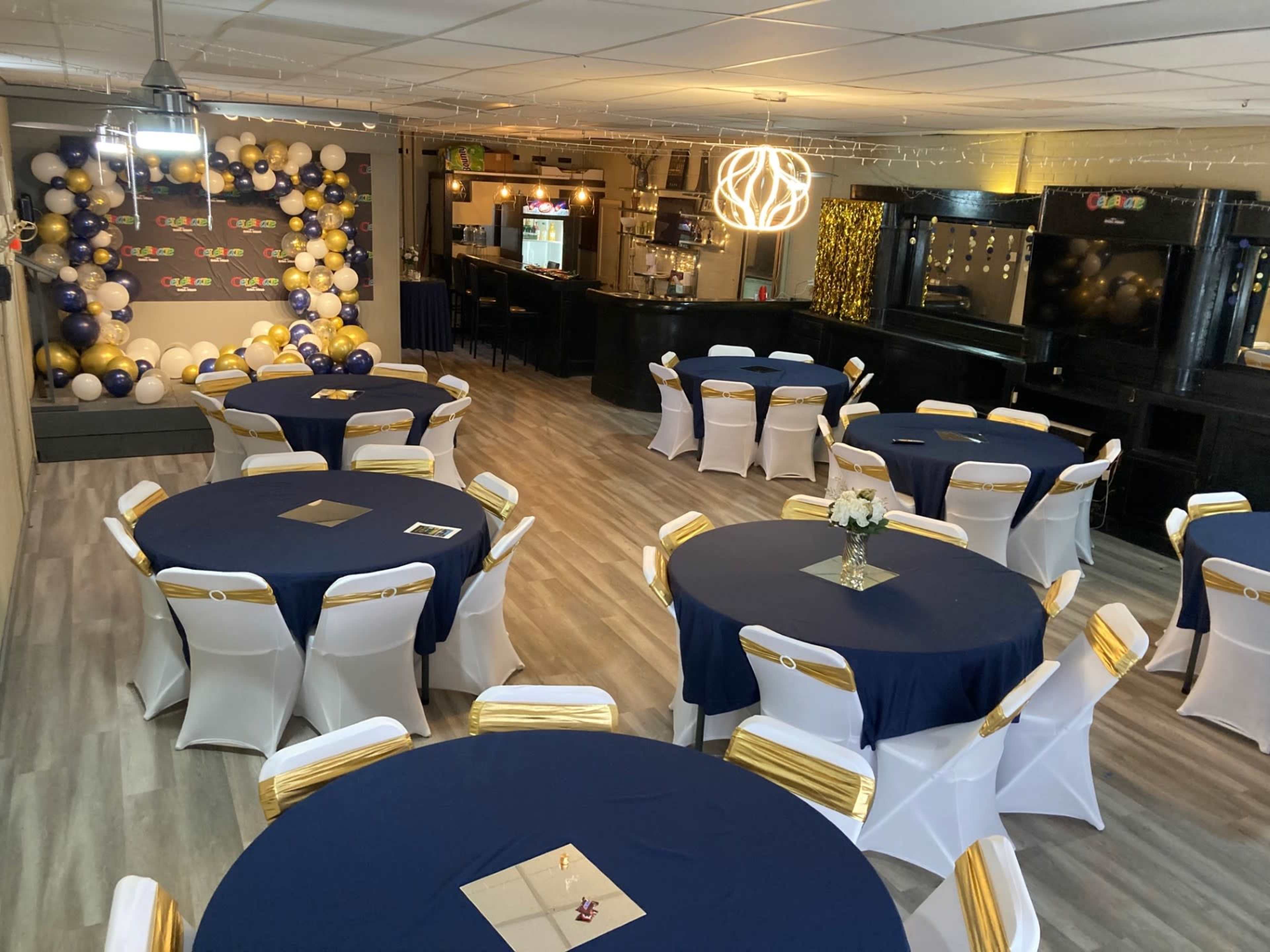 The image shows a room set up for an event with round tables covered in navy tablecloths, white chairs with golden sashes, and a decorative balloon wall in the background.