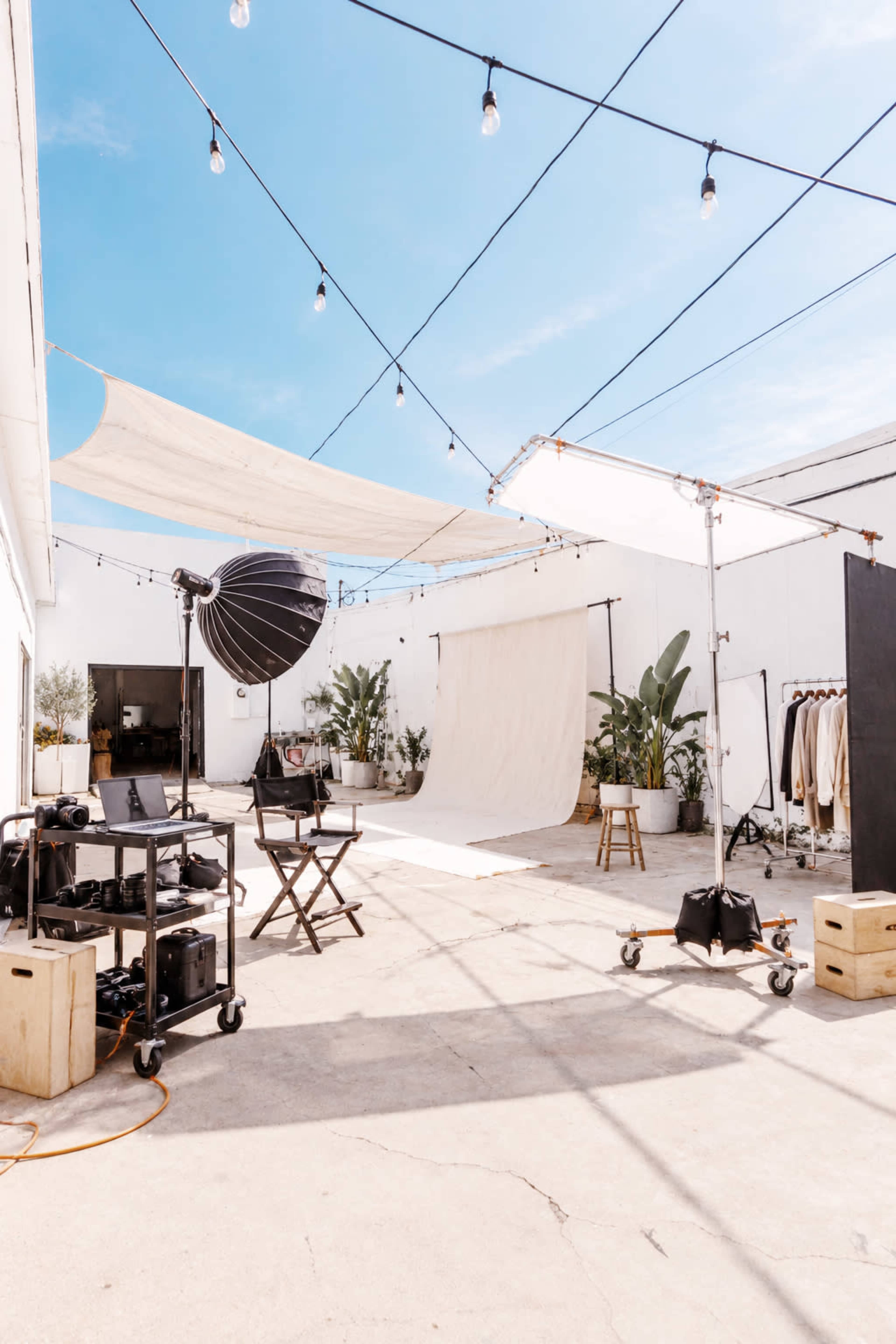 The image shows a spacious photography studio setup outdoors, featuring a backdrop, lighting equipment, and various props surrounded by plants and hanging lights.