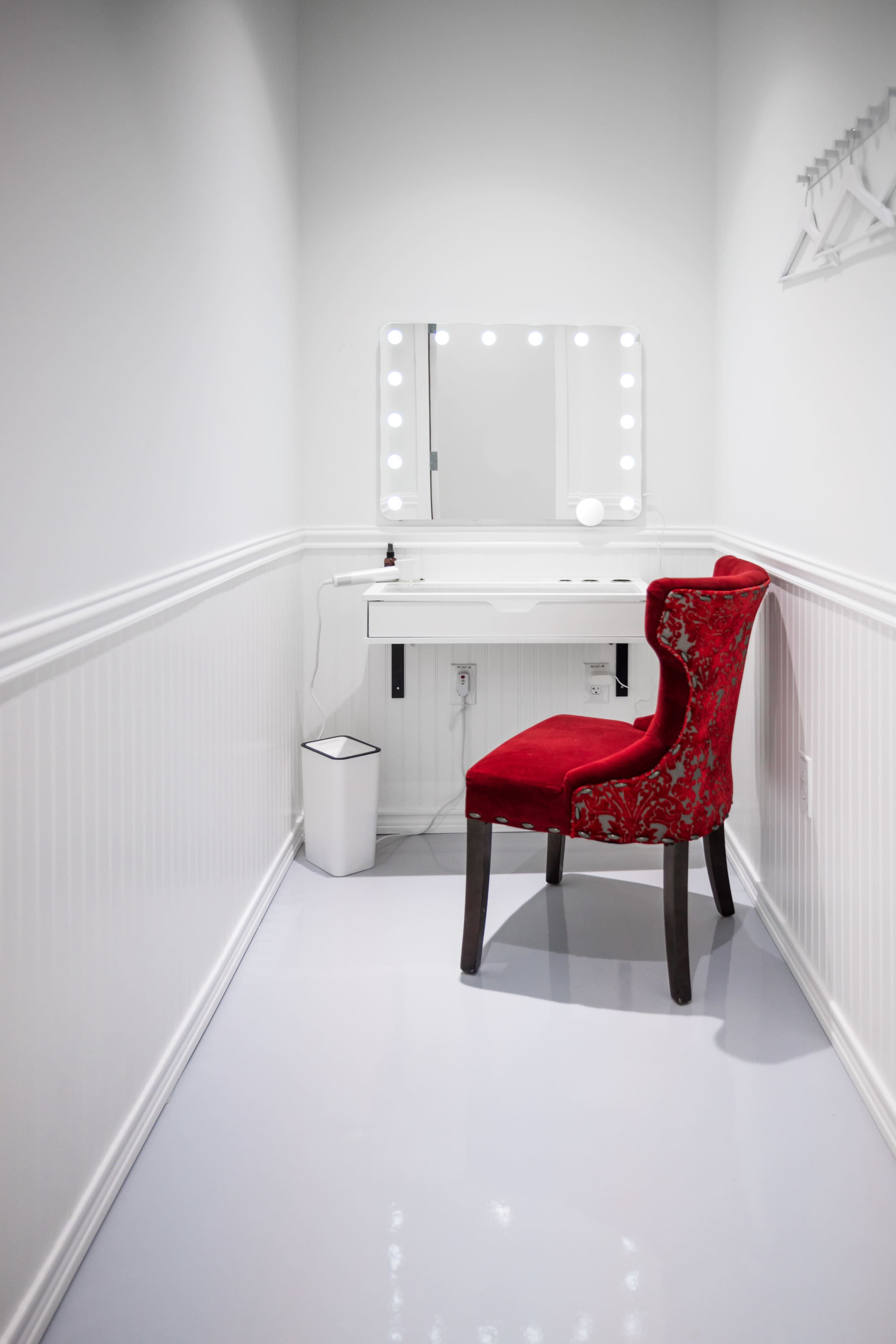 The image shows a narrow, well-lit vanities area featuring a white wall, a small desk with a mirror surrounded by lights, and a red upholstered chair.