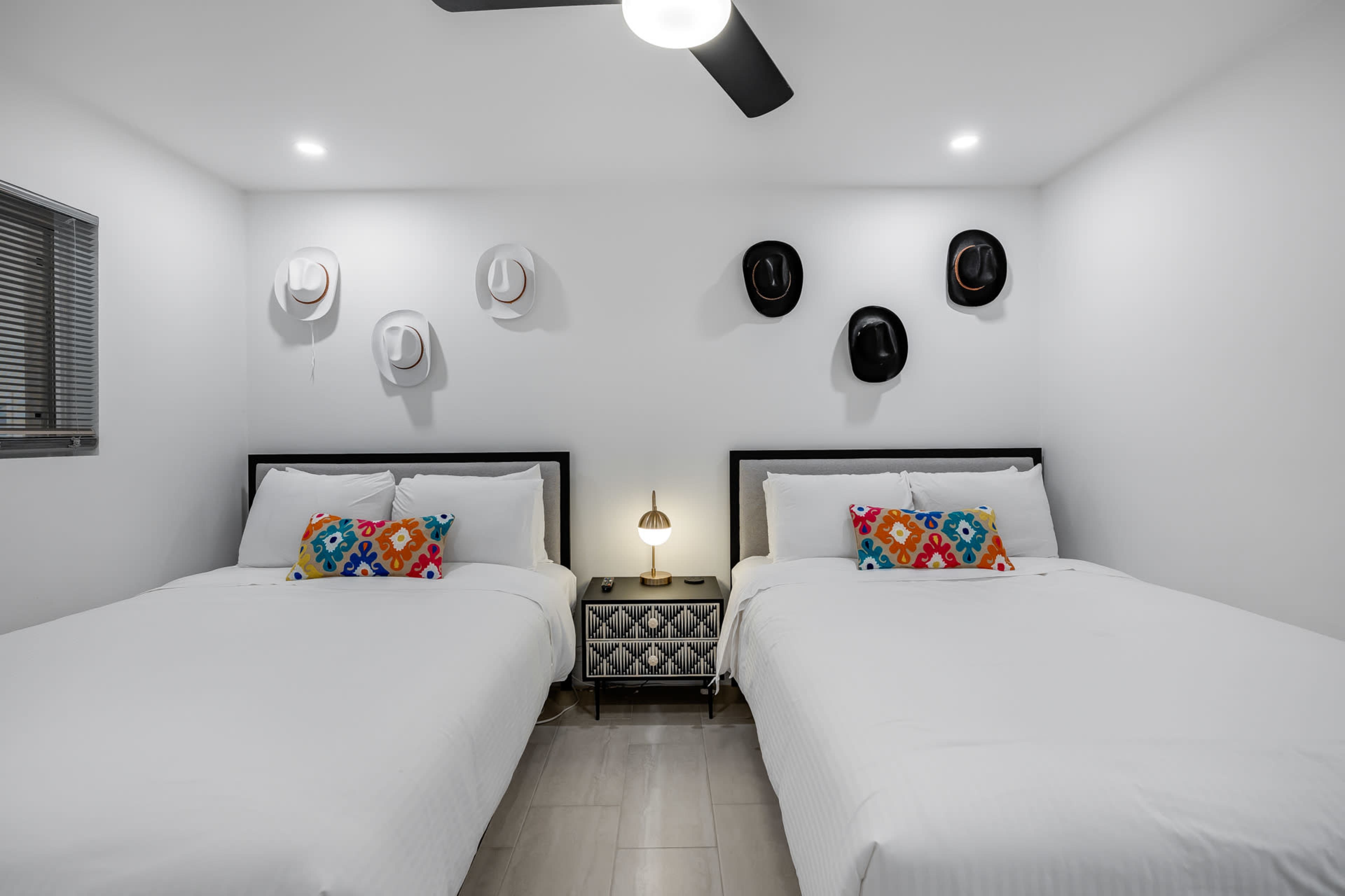 A bedroom with two beds, each dressed in white linens, a bedside table with a lamp, and several hats displayed on the wall.