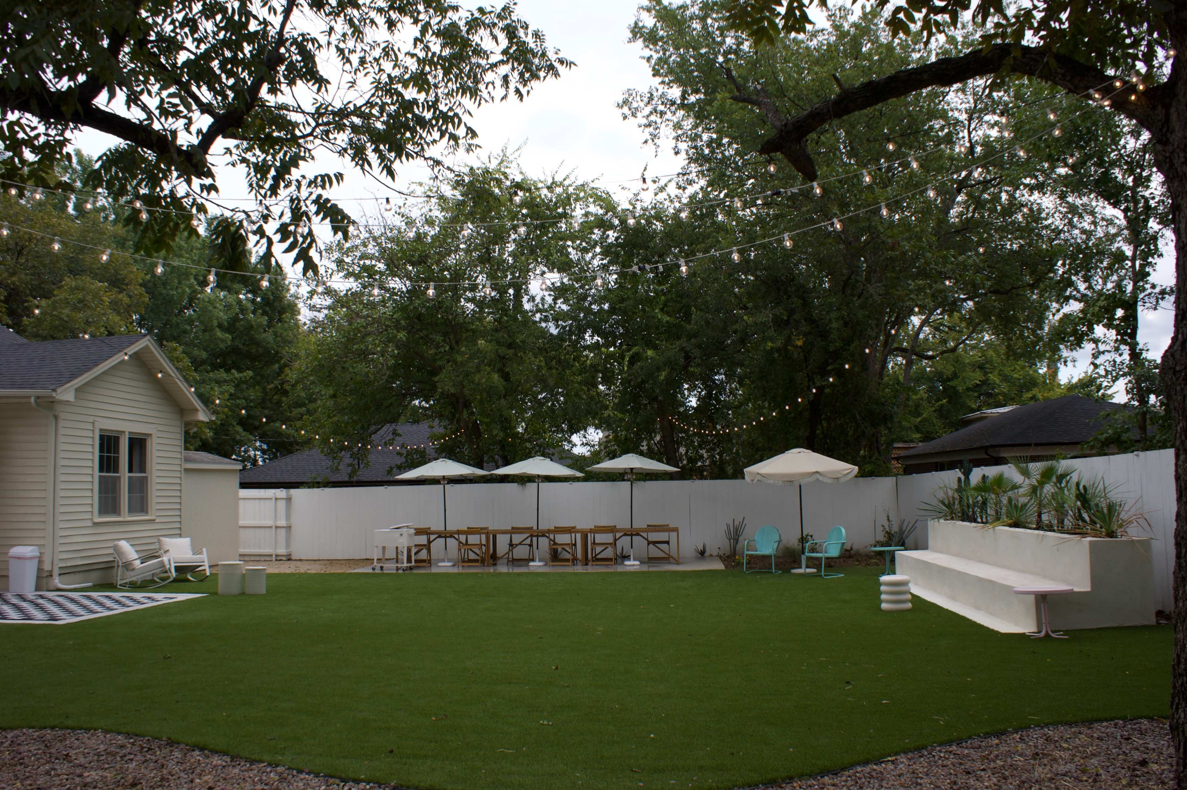 The image features a spacious backyard with artificial turf, a patio area with a picnic table, four umbrellas, and a raised seating area against a white fence, surrounded by trees.