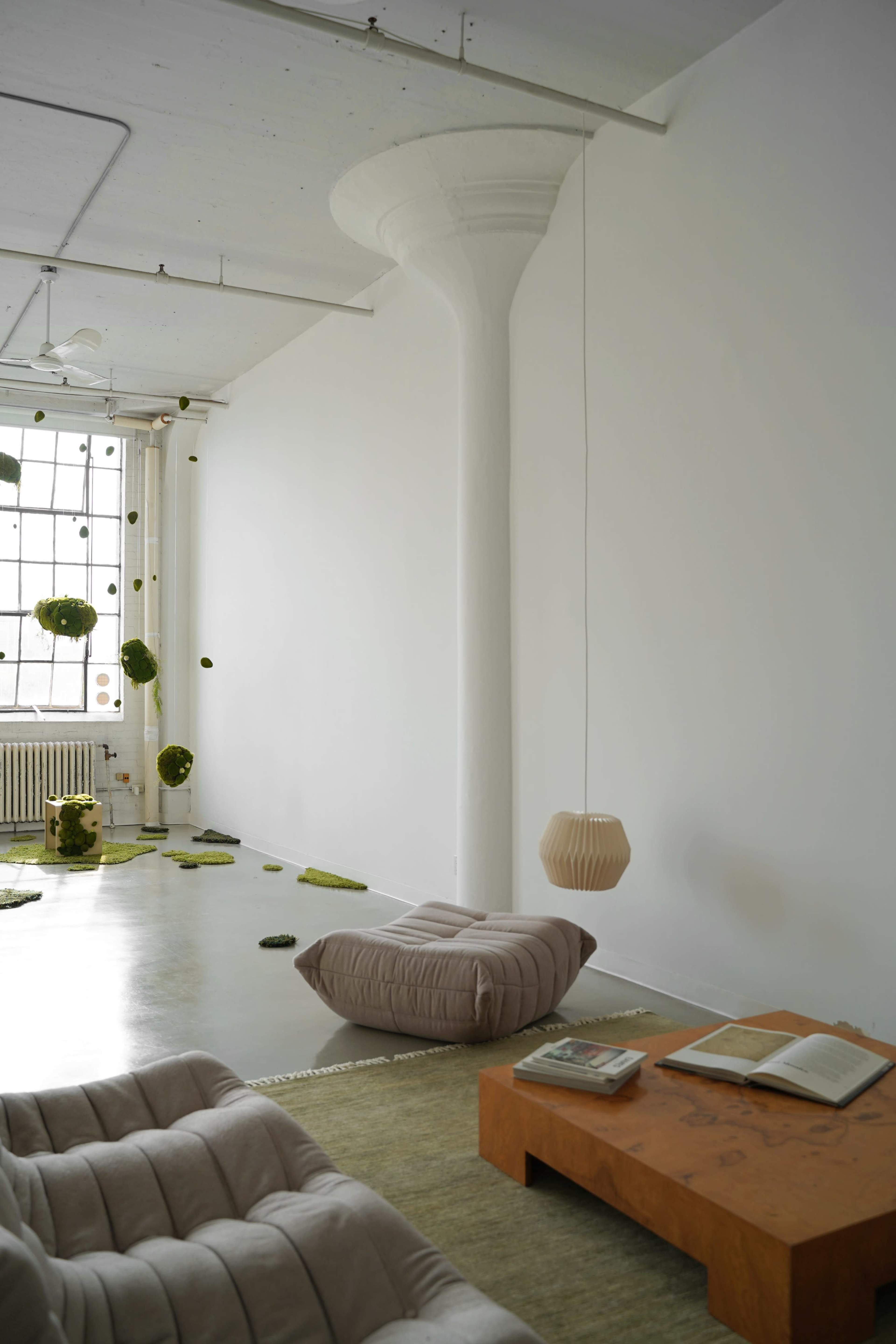 The image shows a minimalistic interior with a white wall, a cozy seating area, a wood table with an open book, and suspended decorative elements.