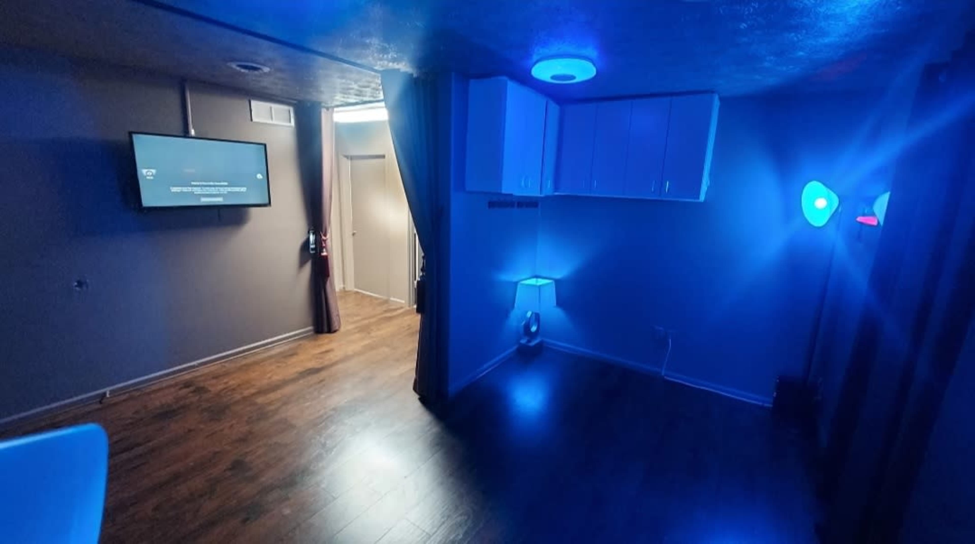 The room features a television mounted on the wall, illuminated by blue ambient lighting, and includes wooden flooring and a lamp.