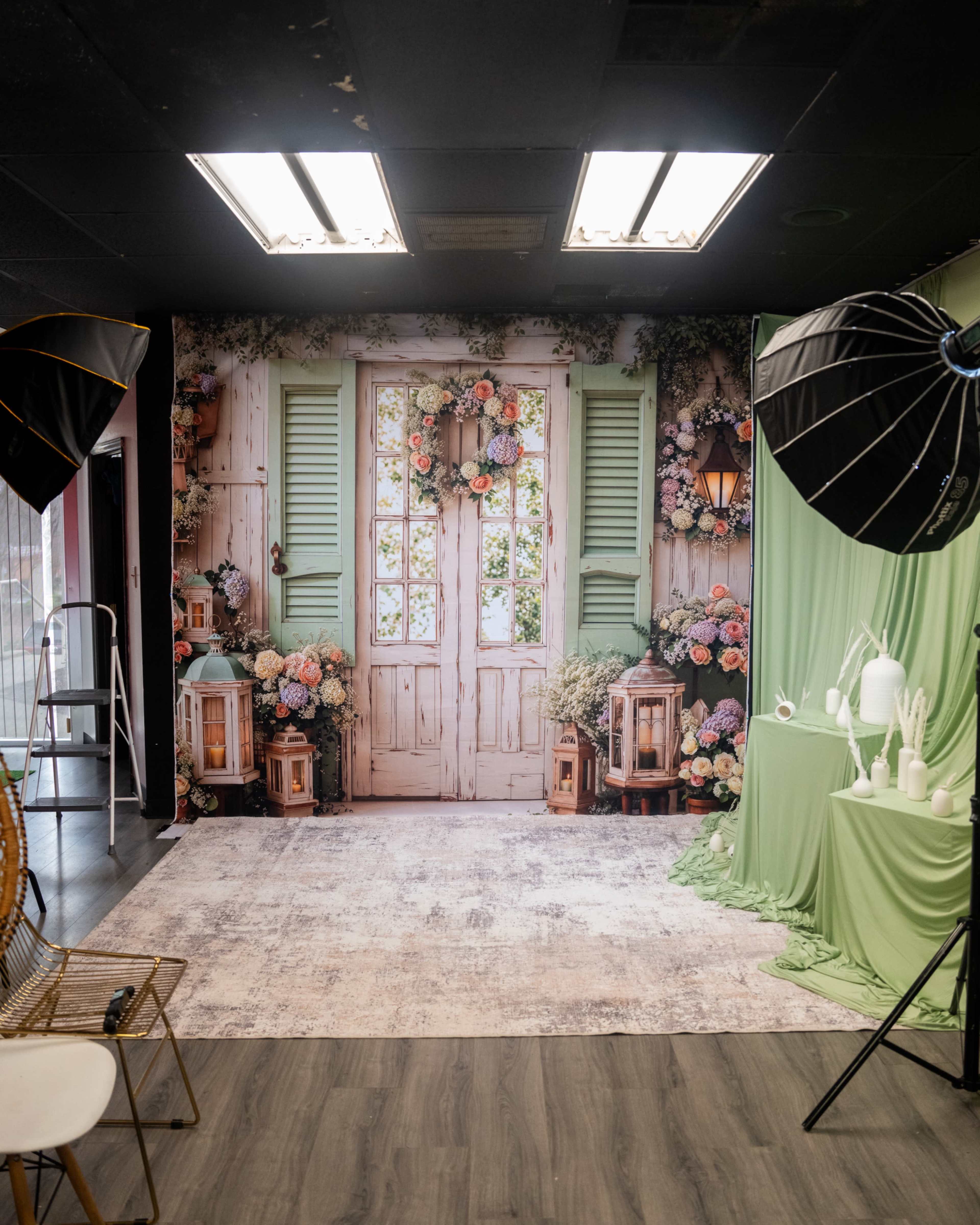 The image shows a photography studio setup featuring a decorative backdrop of a floral-adorned door, flanked by lanterns and green drapery, with studio lighting equipment positioned around the scene.