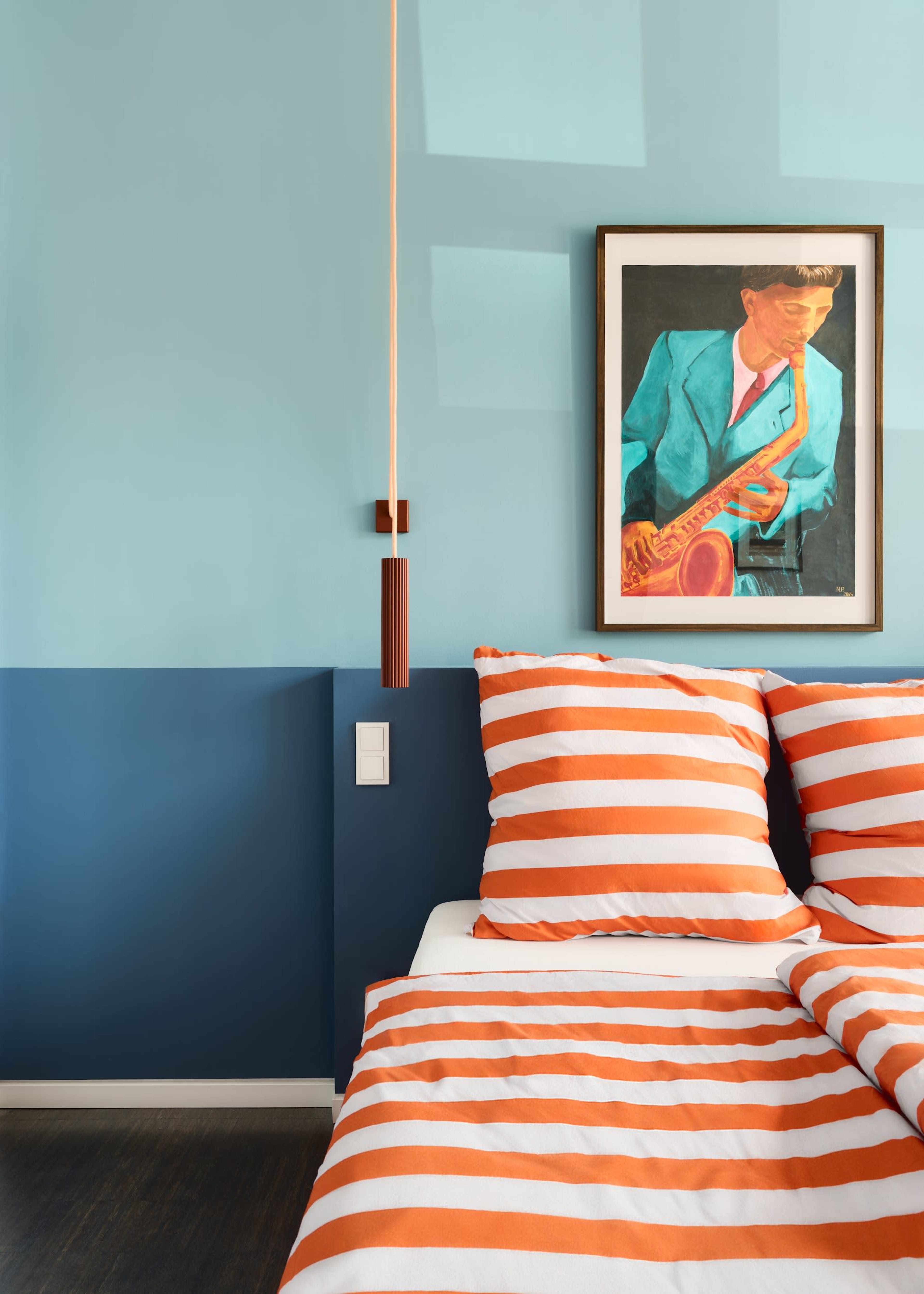 The image shows a modern bedroom with blue and light blue walls, an orange pendant light, striped orange and white bed linens, and a framed portrait of a musician on the wall.