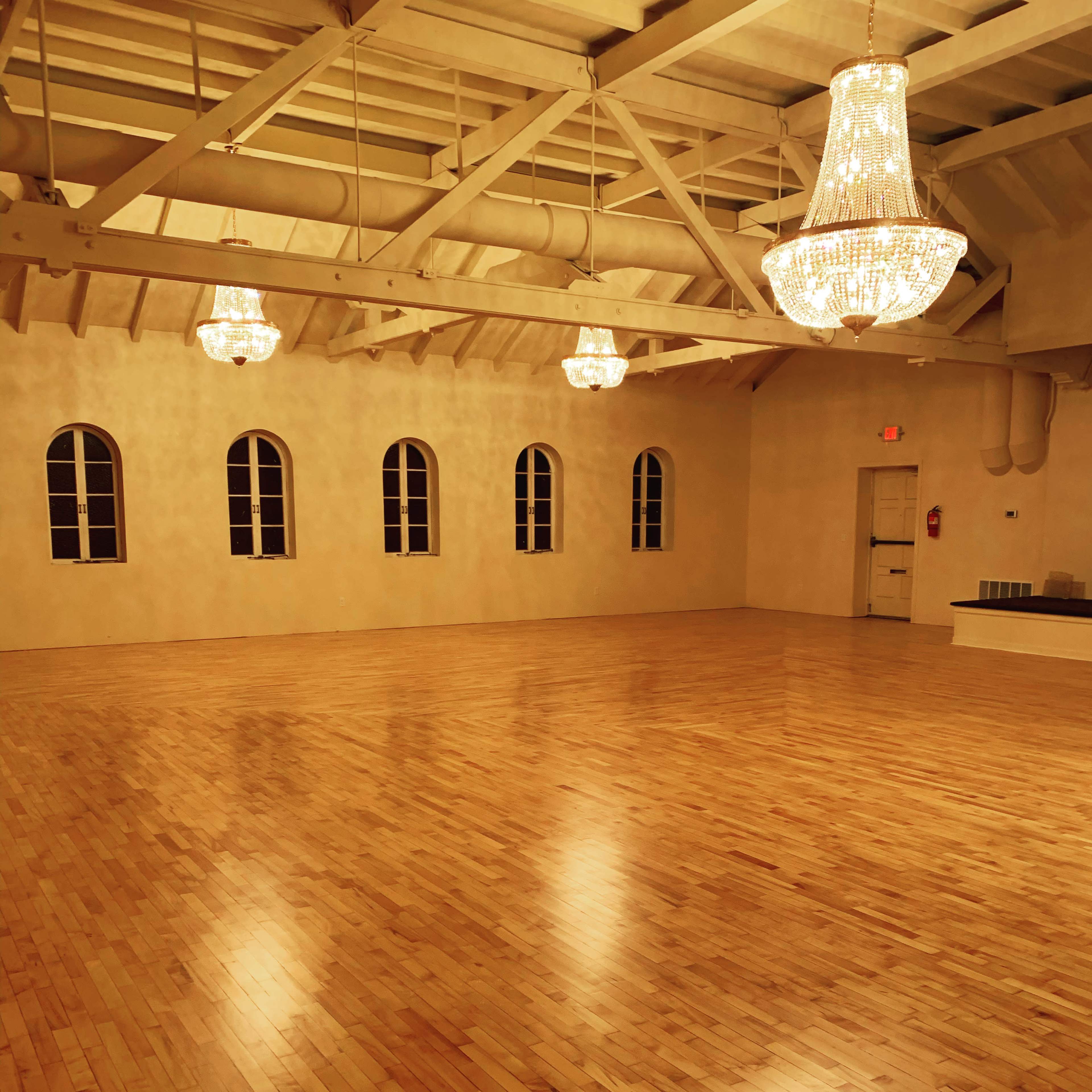 A spacious, empty room with hardwood flooring, three chandeliers hanging from the ceiling, and several arched windows along the walls.