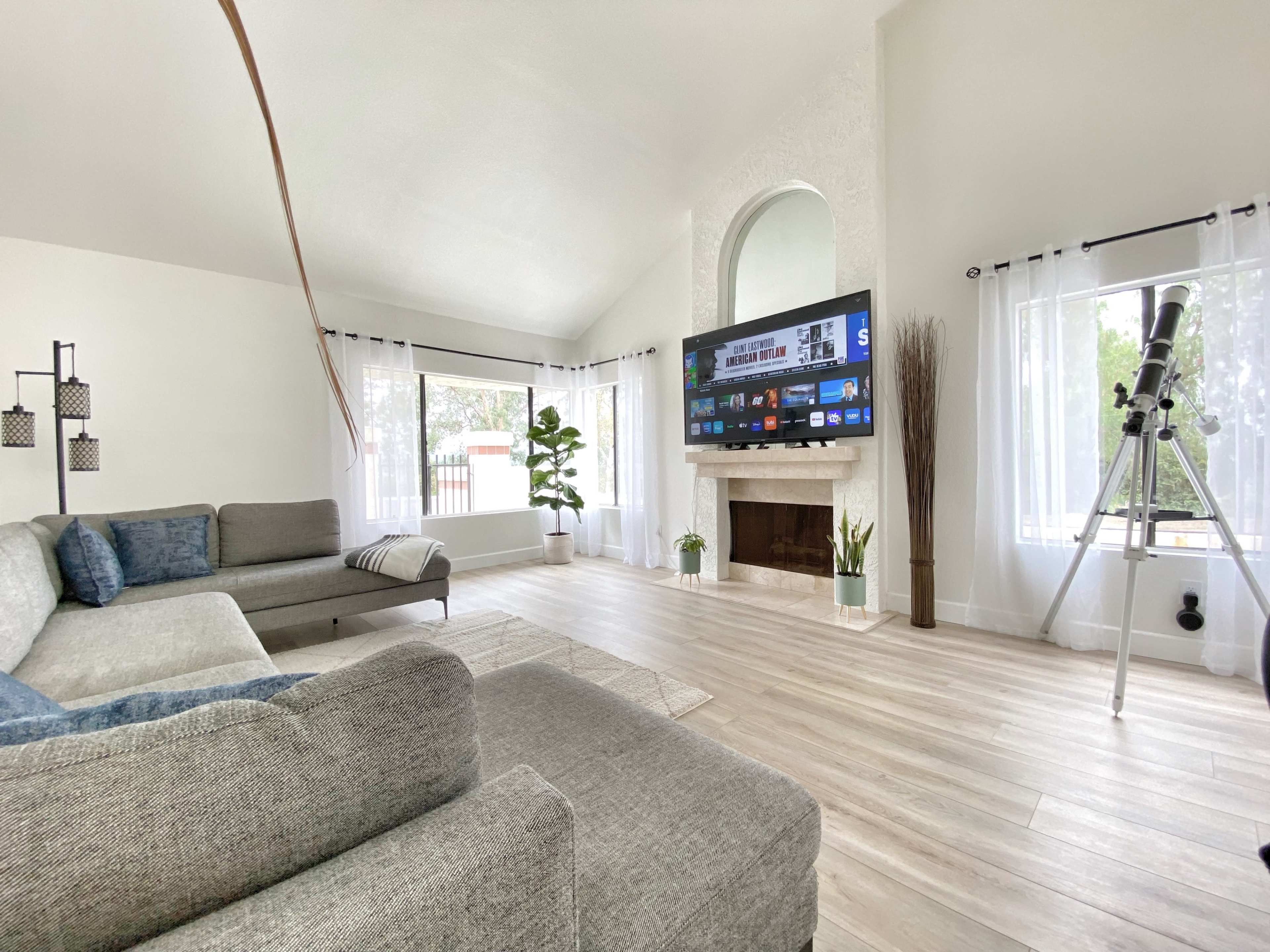 The bright living room features a large television mounted on the wall opposite a gray sectional sofa, with a telescope positioned near the window.
