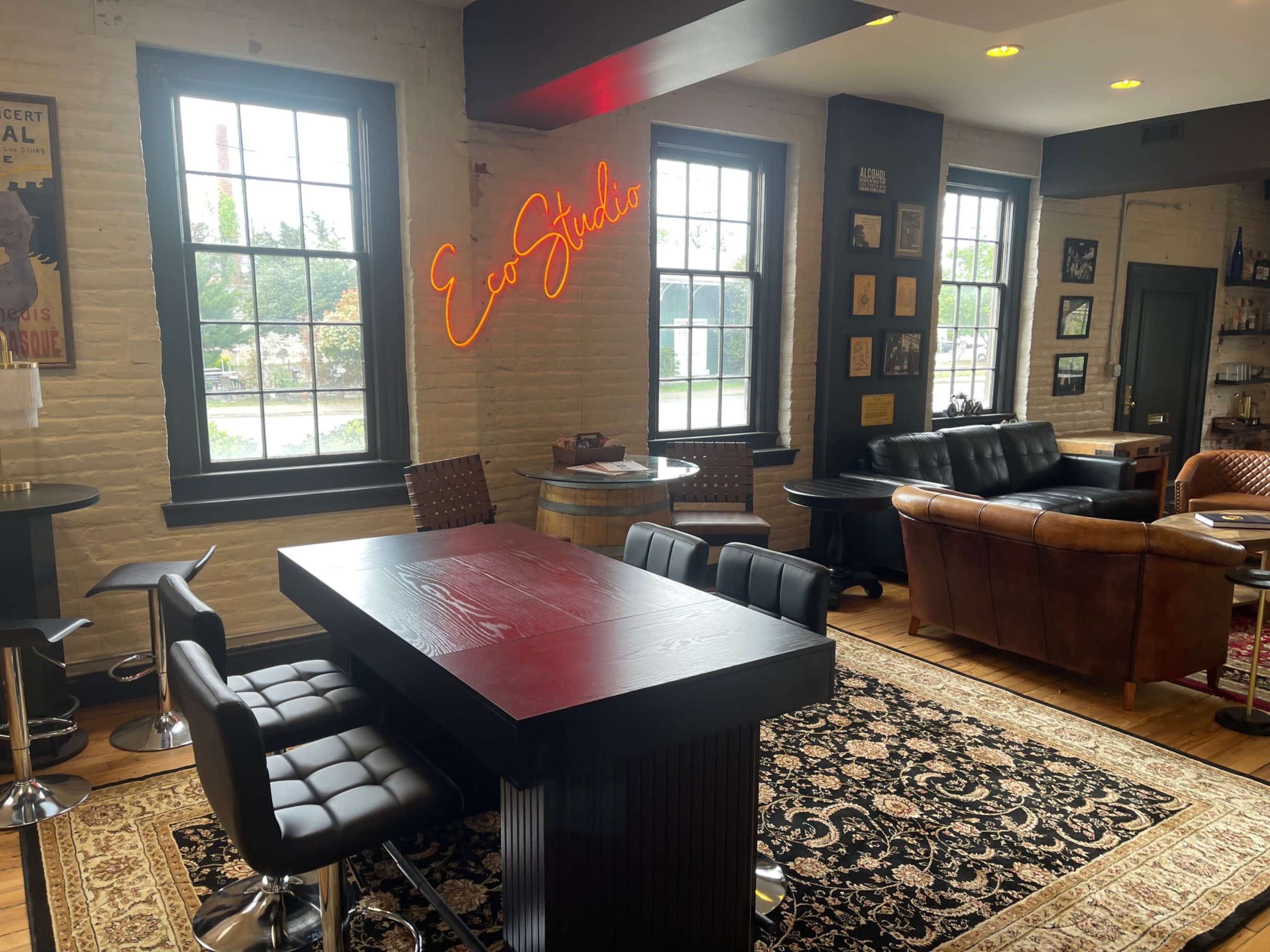 The image shows a cozy interior space that features a dark wood table with bar stools, vintage decor, and a neon sign reading "Eco Studio."