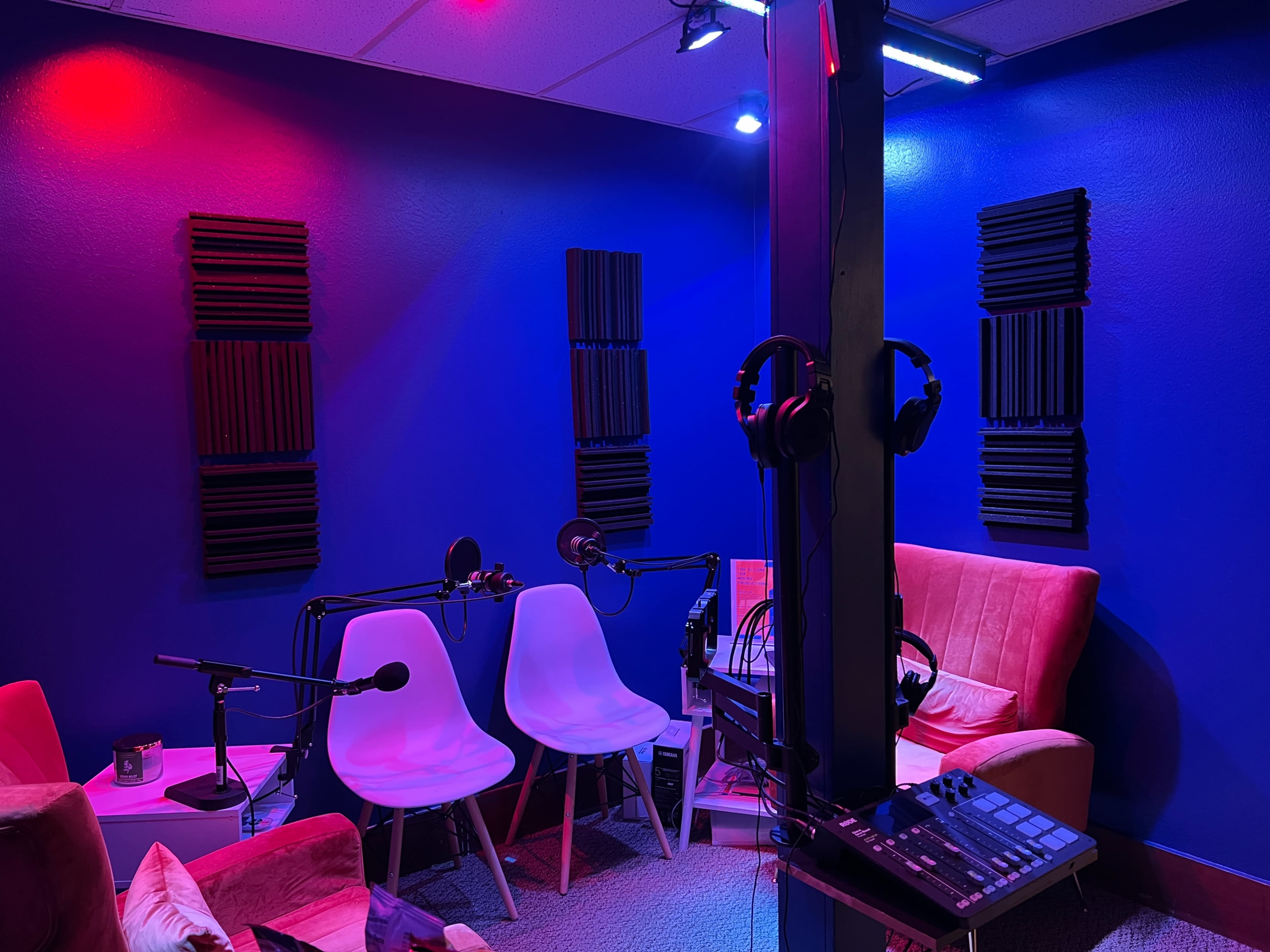 A small recording studio with two white chairs, microphones on stands, soundproofing panels on the walls, and a mixing console.