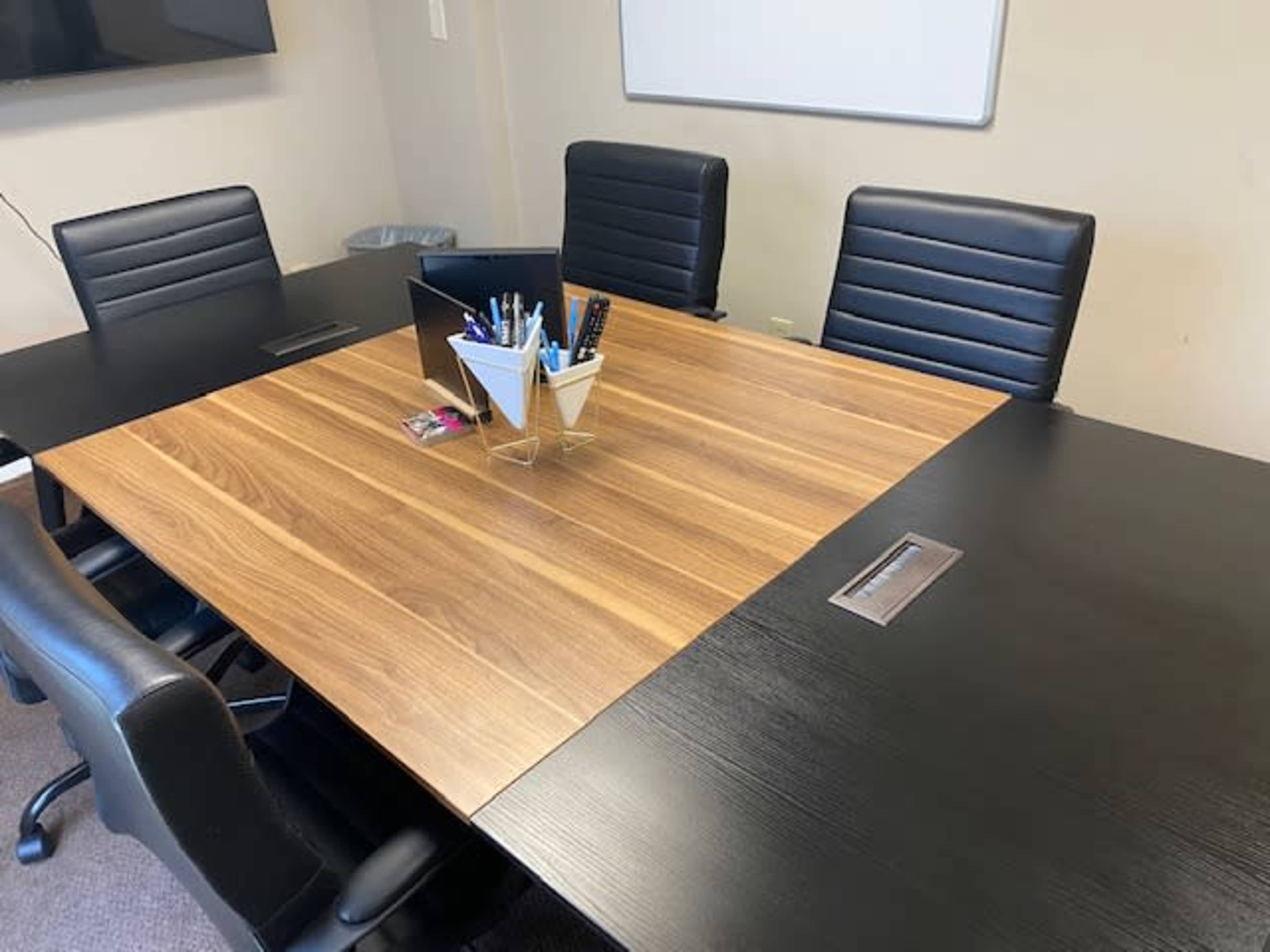 The image shows a conference room table with a wooden top, surrounded by six black leather chairs, and a container of pens and stationery in the center.