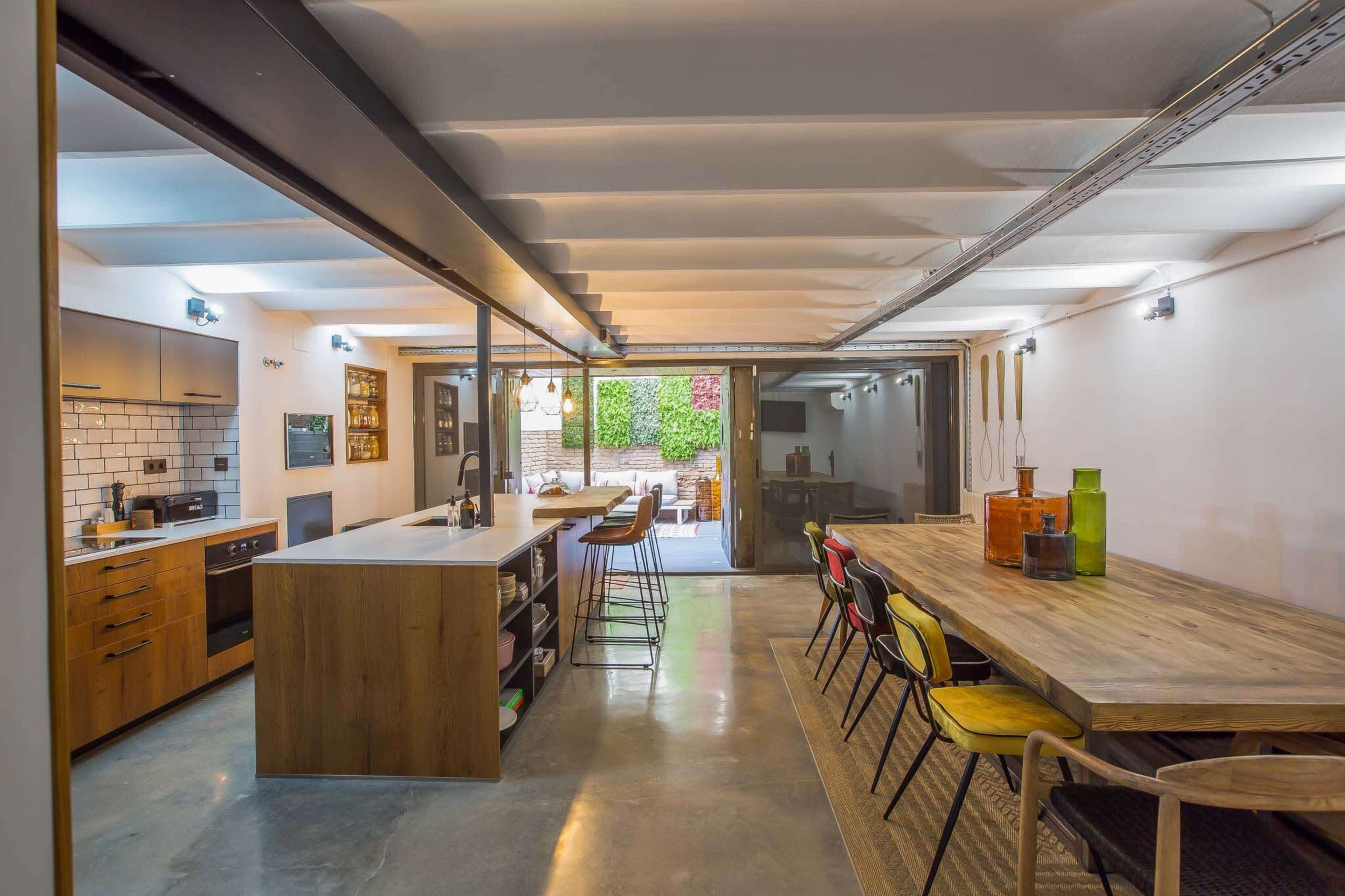 A modern kitchen and dining area with wooden furnishings, concrete flooring, and large glass doors opening to an outdoor space.