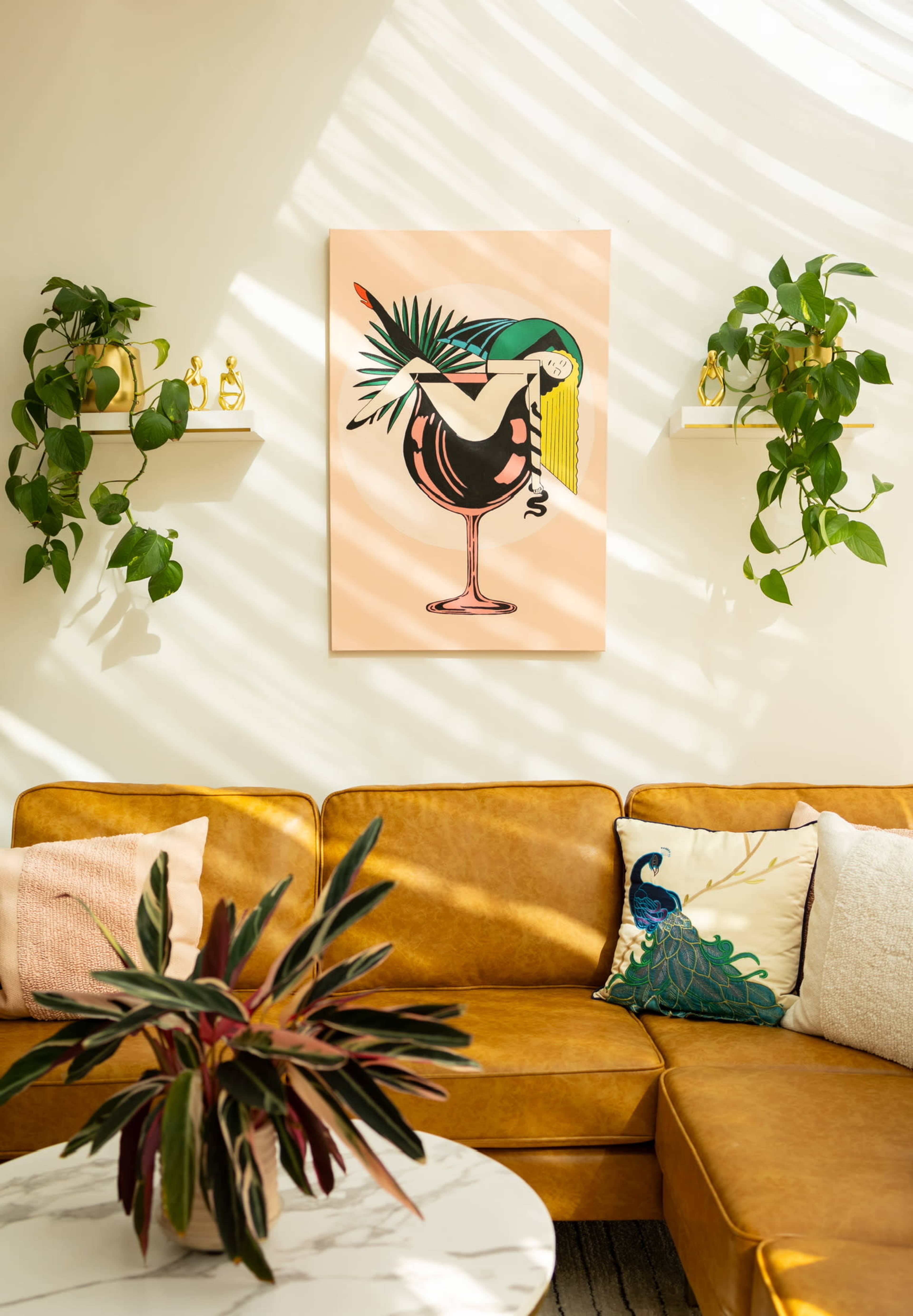 The image shows a living room with a brown leather couch, a vibrant artwork featuring a bird and cocktail, and green plants on shelves.