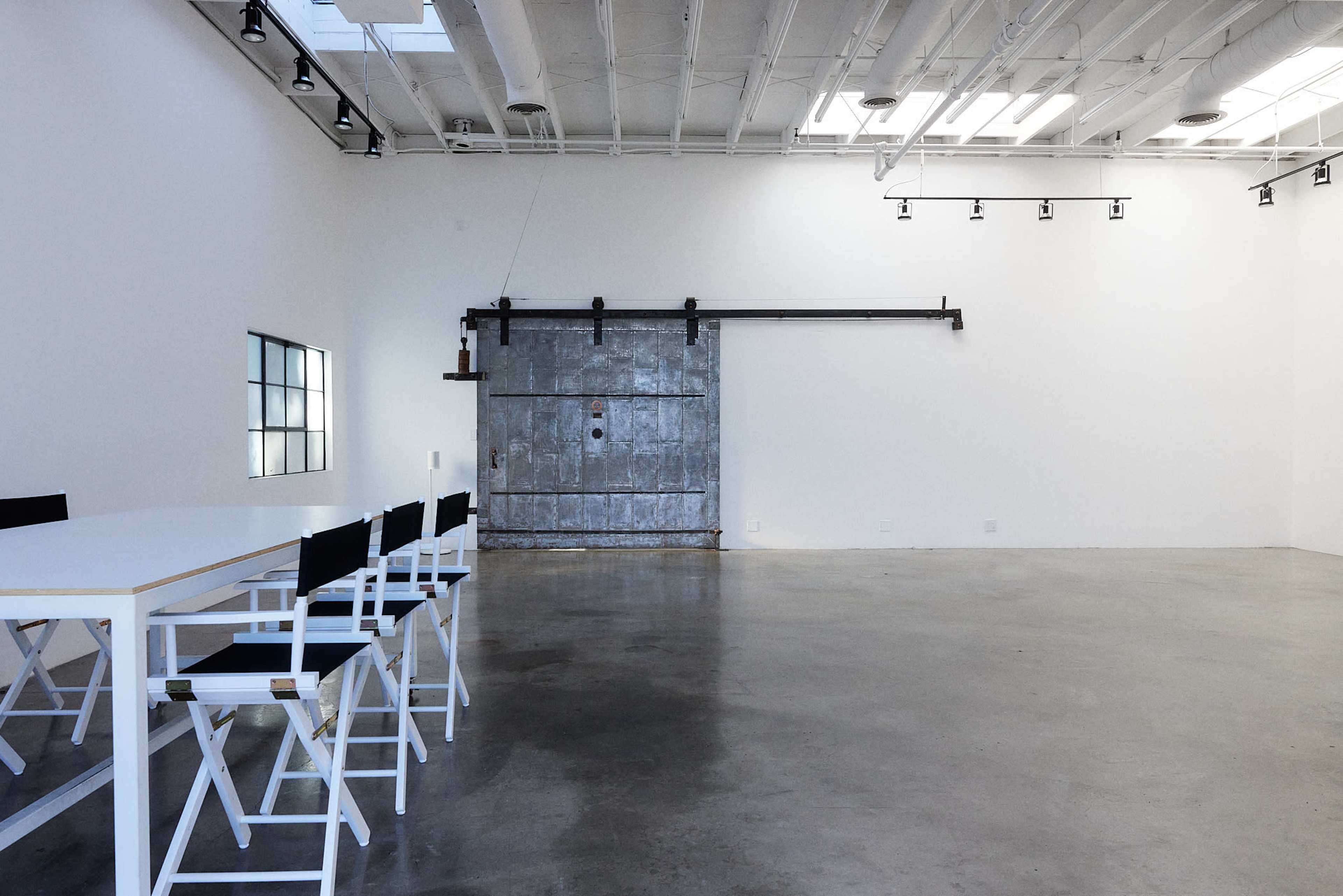 A spacious, minimalist indoor area with a long white table and several folding chairs facing a large metal sliding door.
