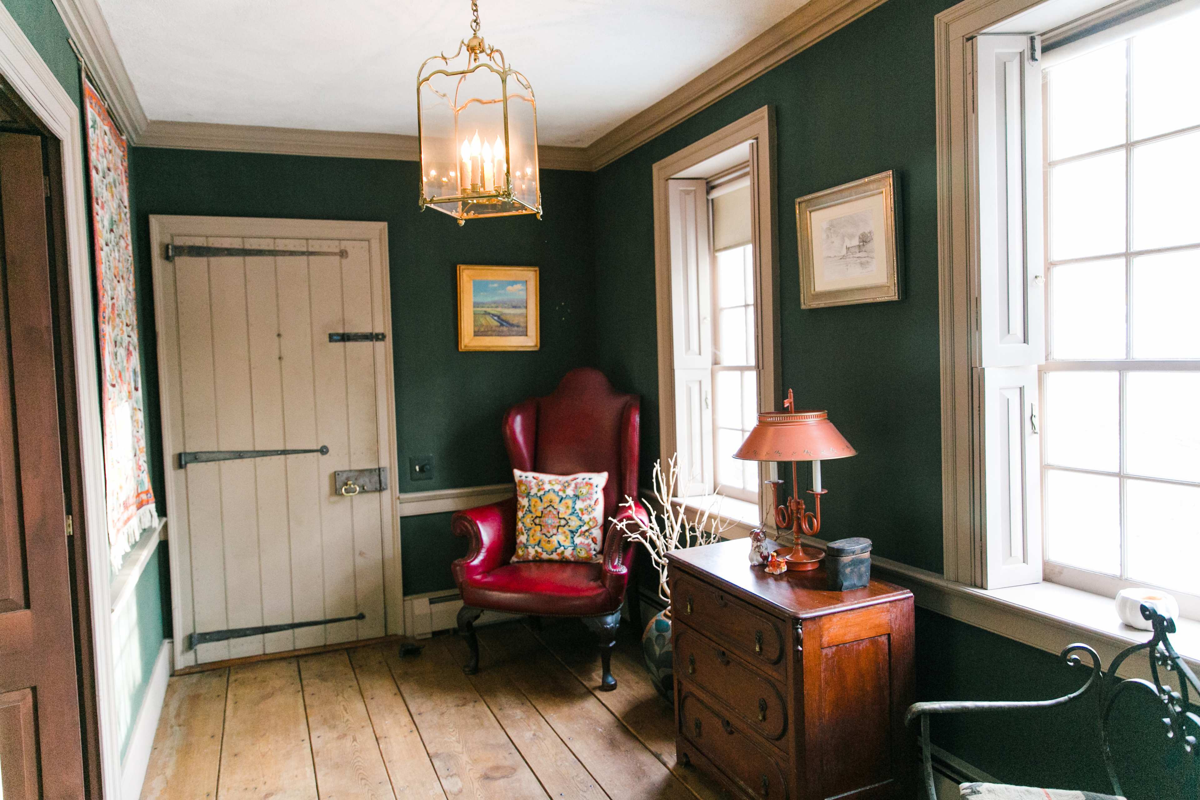 A red velvet armchair is positioned beside a wooden chest of drawers in a room with dark green walls and two windows allowing natural light.