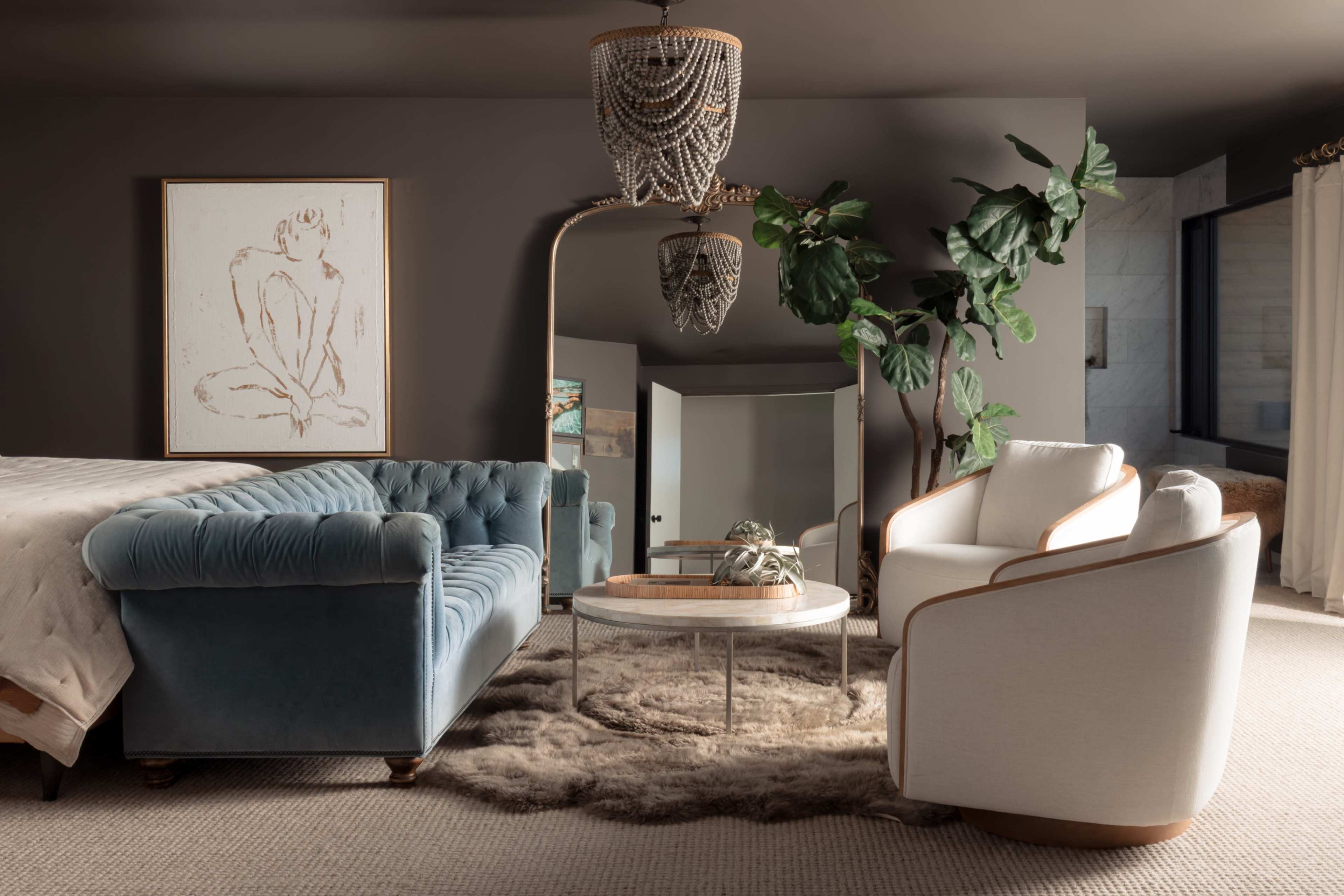 The image shows a stylish living area featuring a blue tufted sofa, a white armchair, a round glass coffee table, and a large potted plant, all set against a dark wall with a piece of art and a decorative mirror.