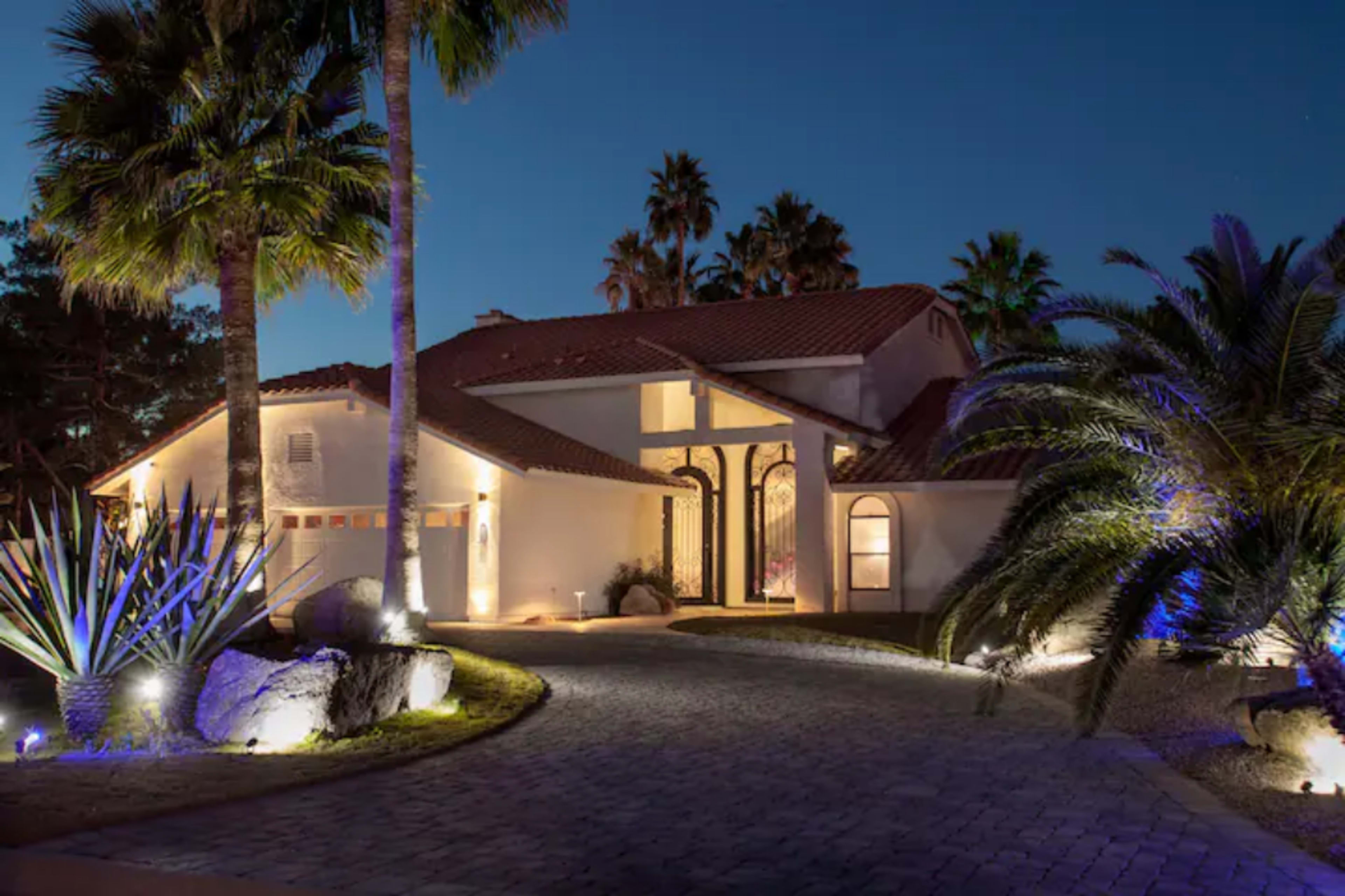 A large, well-lit house with a red-tiled roof is situated amidst palm trees and landscaped garden features under a night sky.