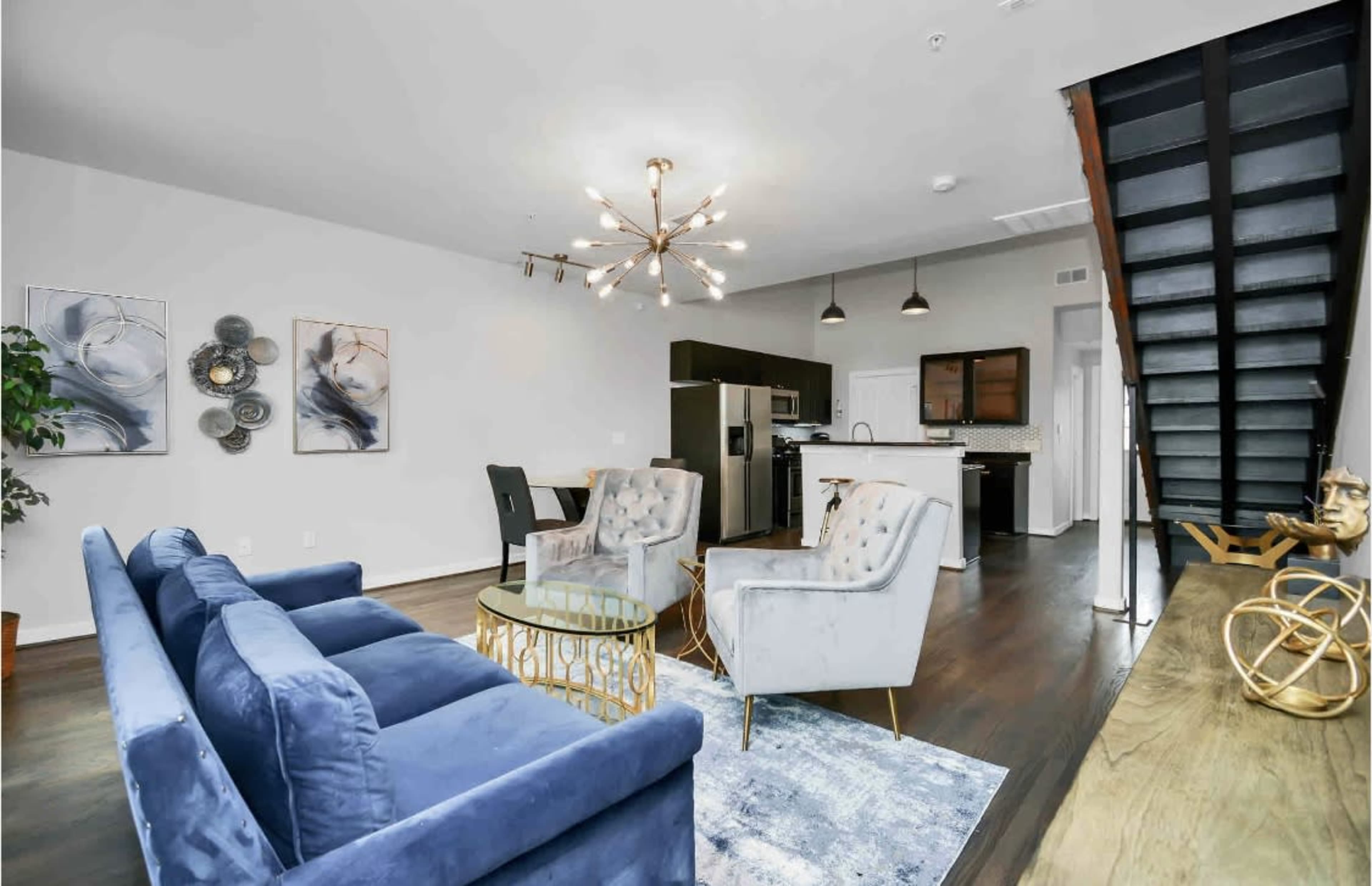 The image shows a modern living space with a blue velvet sofa, a round coffee table, a dining area, and a staircase leading to an upper level.