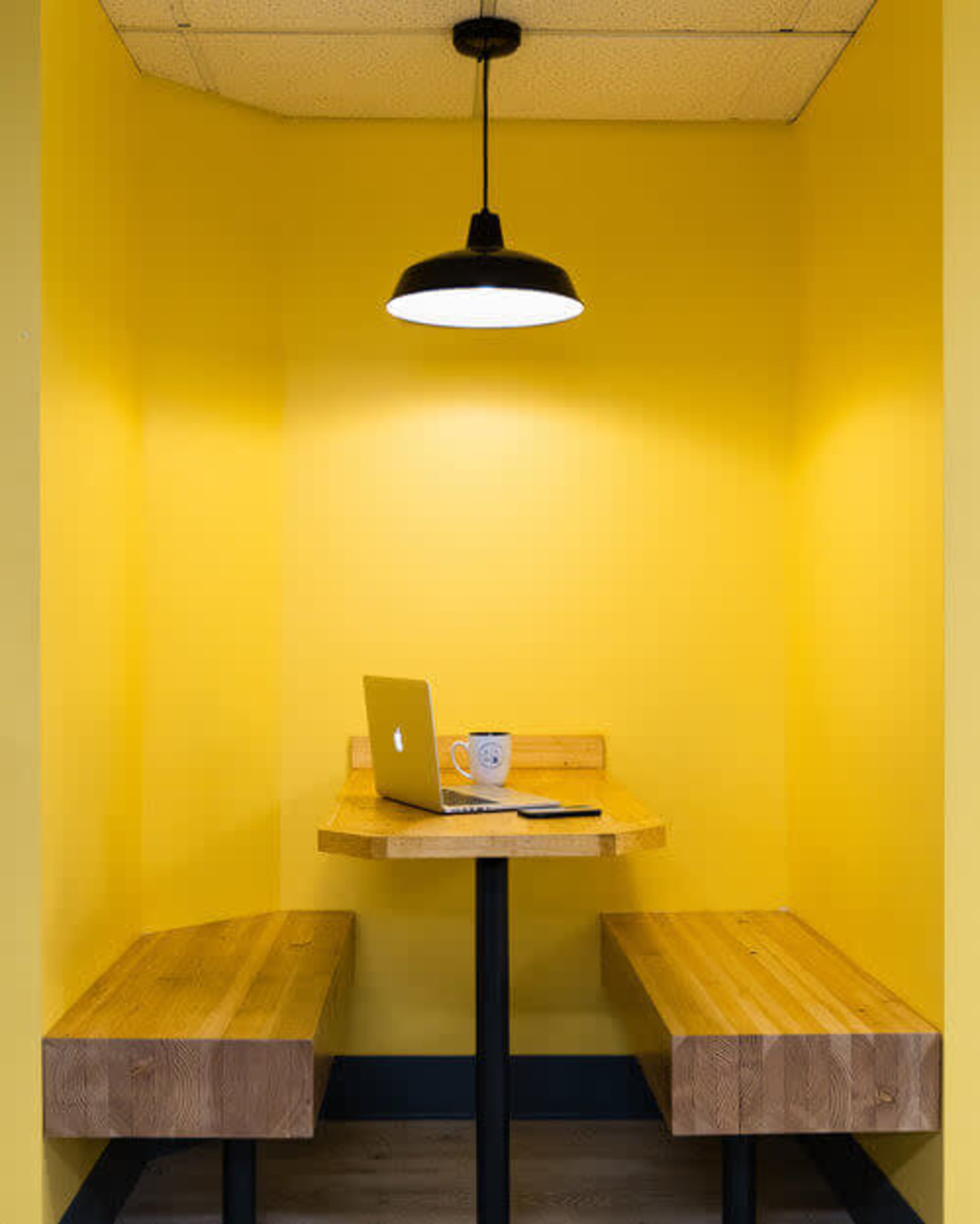 The image shows a small, bright yellow nook featuring a wooden table with two built-in benches and a laptop, accompanied by a coffee mug.