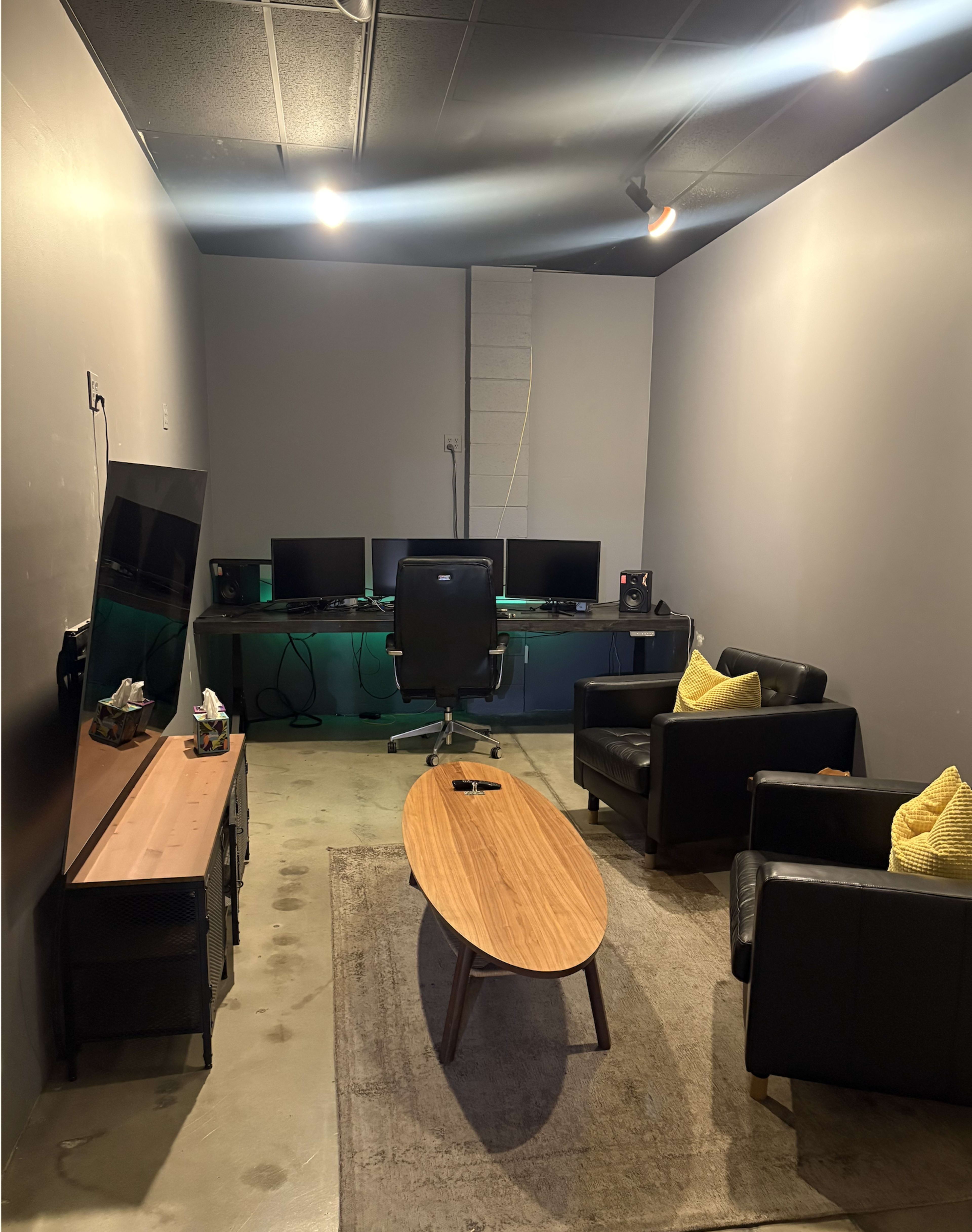 The image shows a modern office space featuring two black leather chairs, a wooden coffee table, a desk with multiple computer monitors, and a television against the wall.