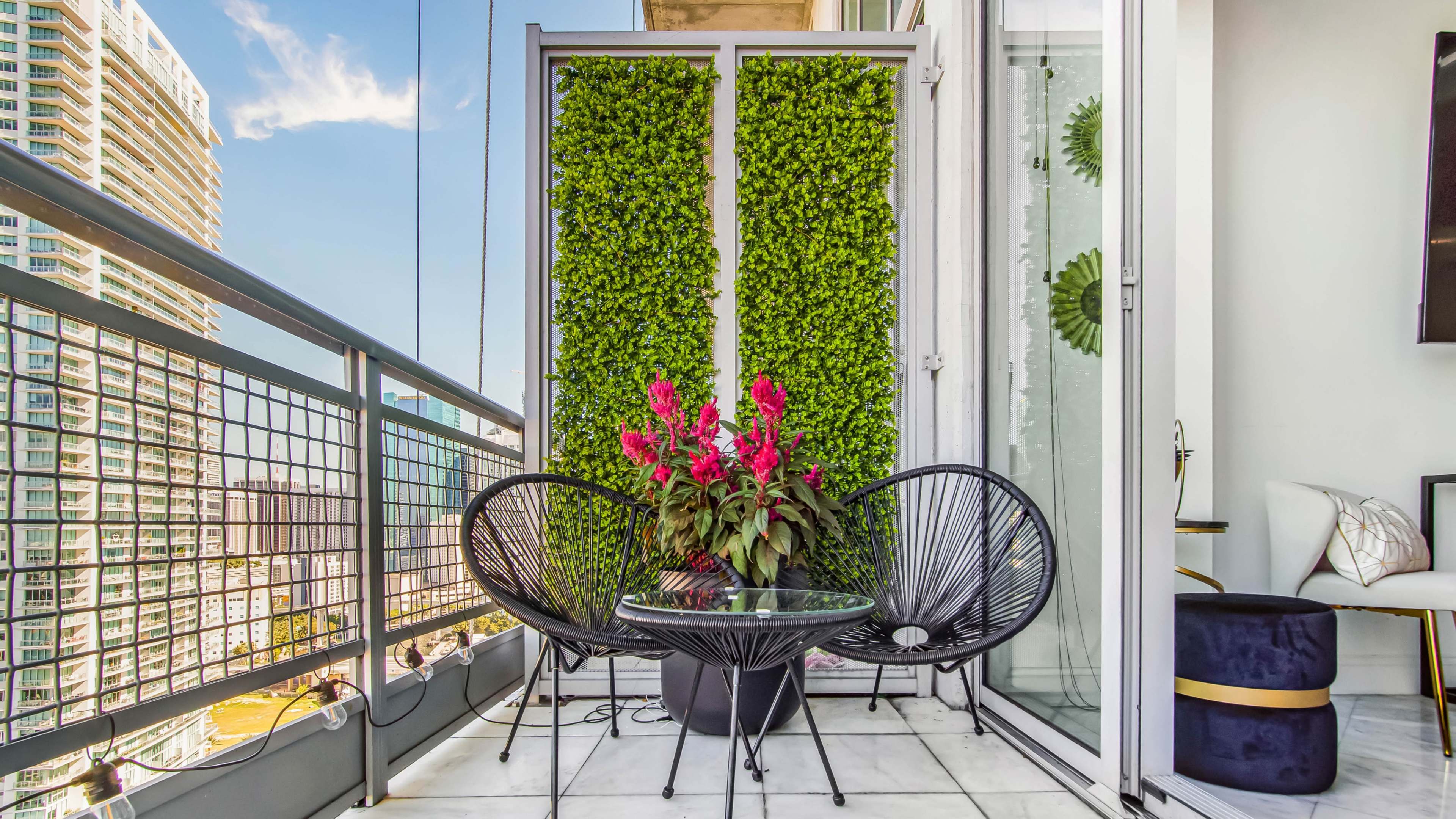 A balcony with two black chairs, a small glass table, and a vertical garden panel with pink flowers, surrounded by a cityscape view.