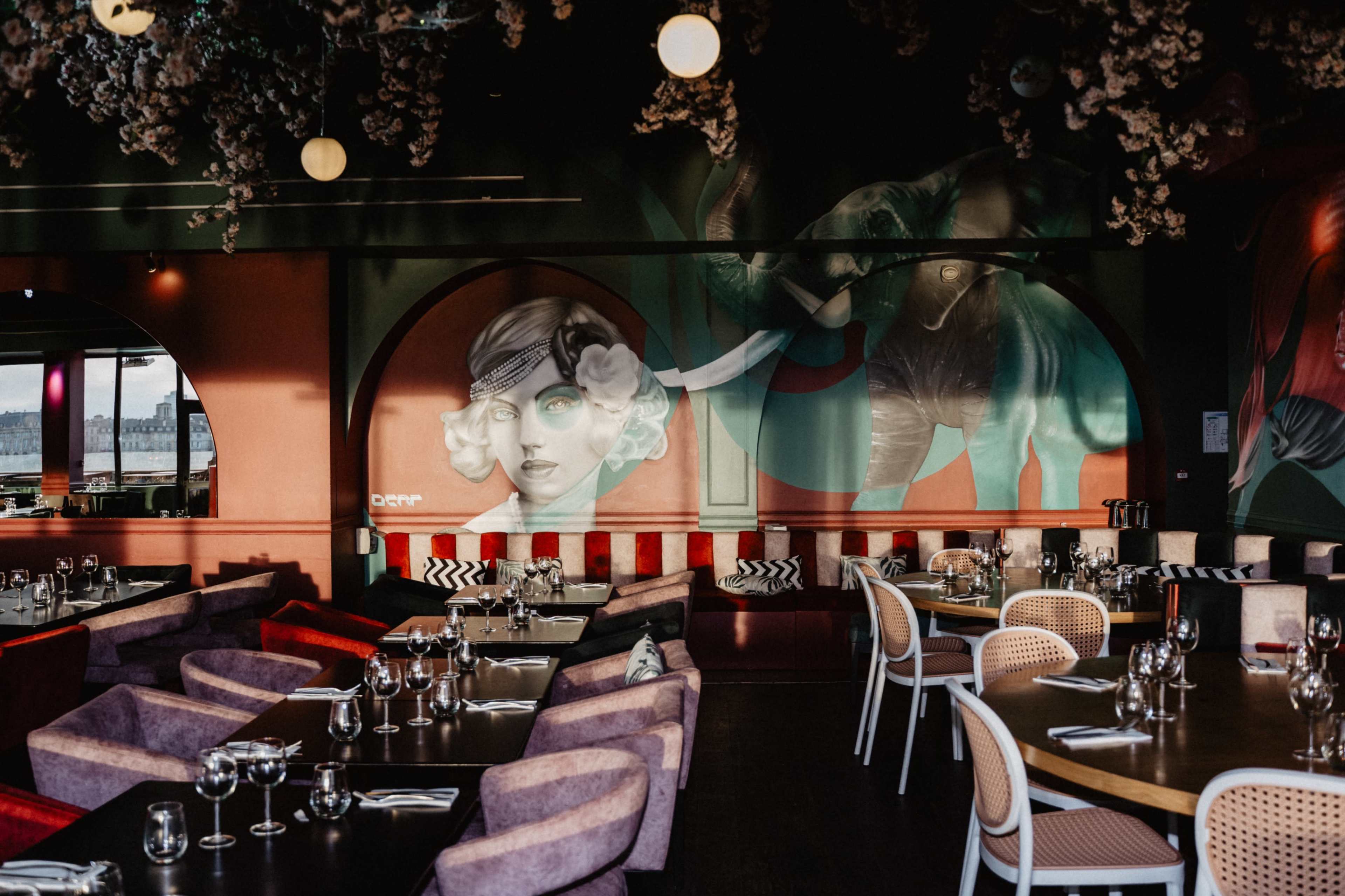The interior of a restaurant features colorful murals, stylish seating, and neatly arranged tables set with glassware.