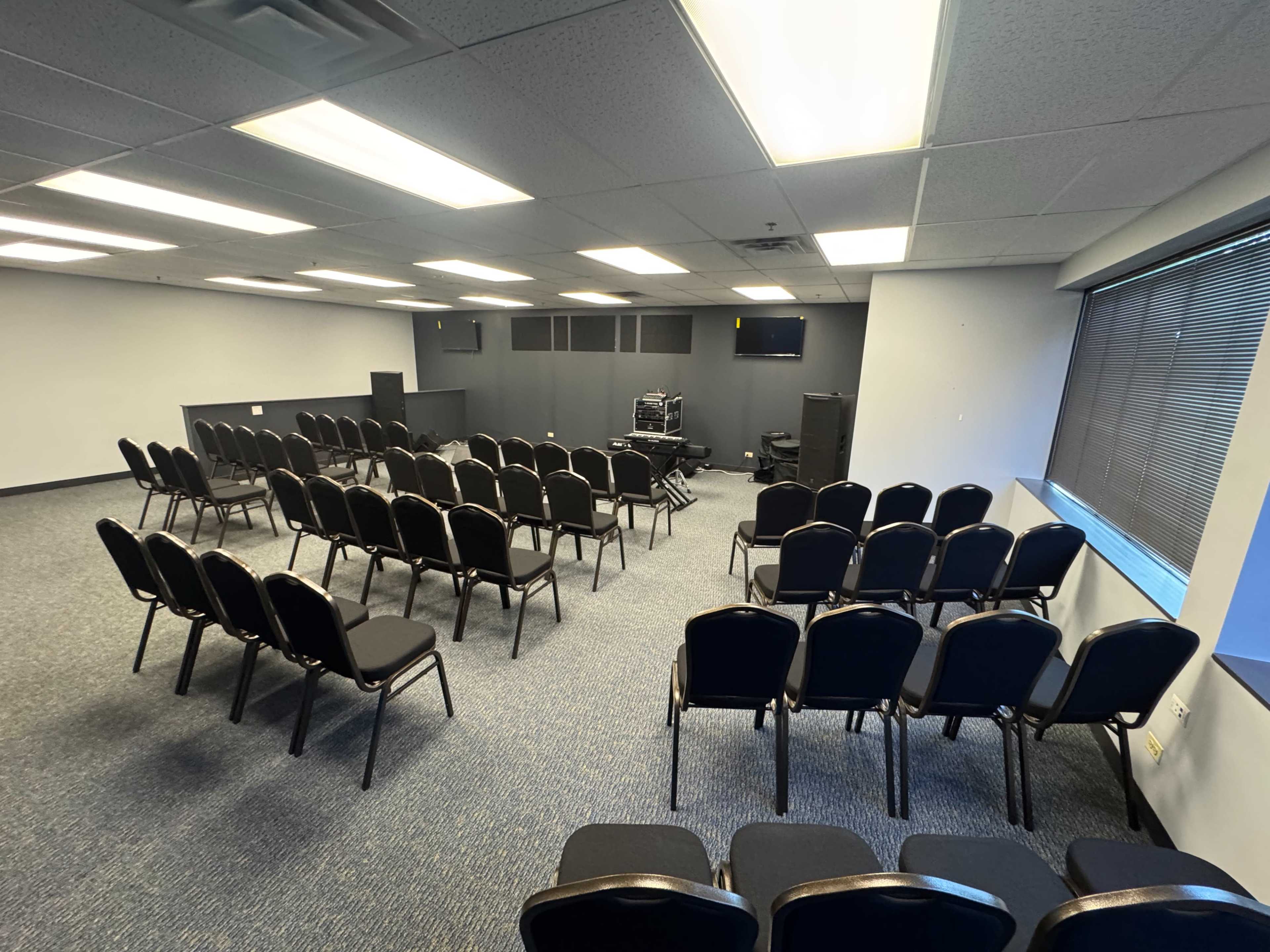 The image shows a conference room with neatly arranged rows of black chairs, a few audio equipment pieces in the back, and windows along one wall.