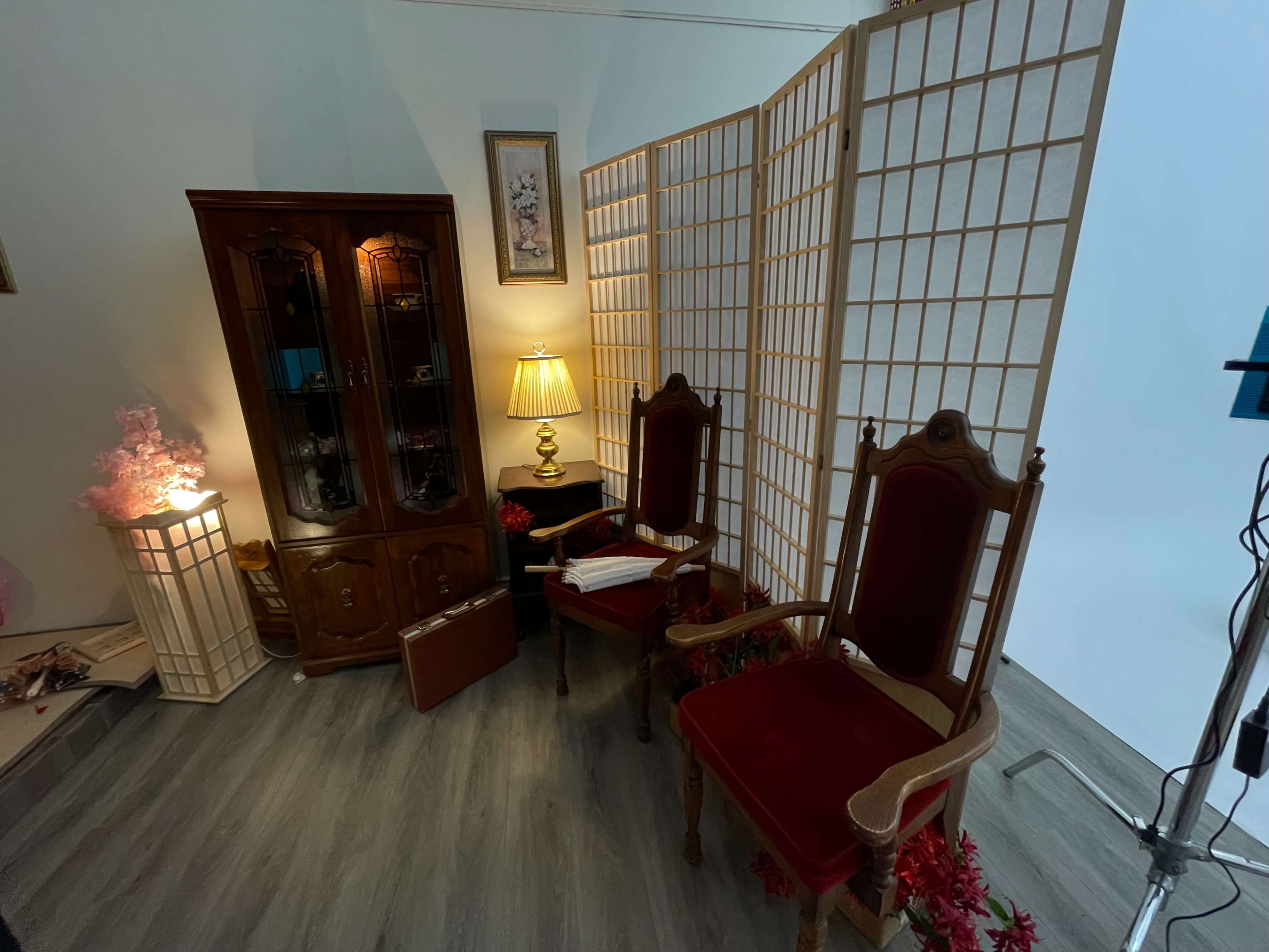 The image shows a small room featuring two ornate wooden chairs, a glass-front cabinet, a lamp, and a folding screen, with decorative accents and plants.