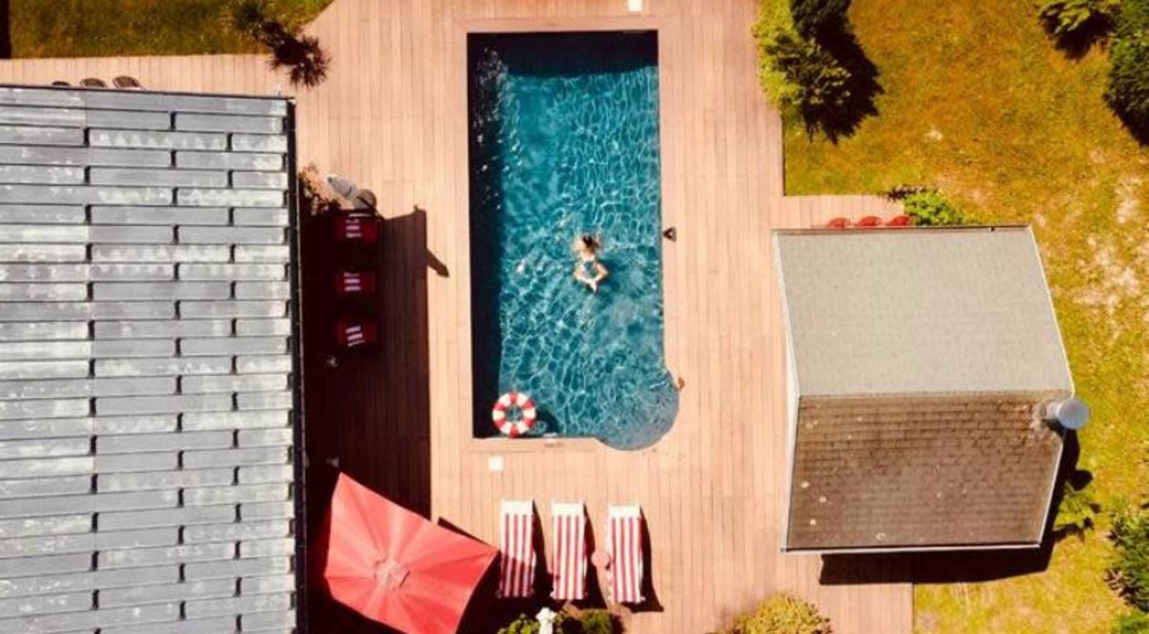 An aerial view shows a rectangular swimming pool surrounded by wooden decking, lounge chairs, and a nearby shed and patio with an umbrella.