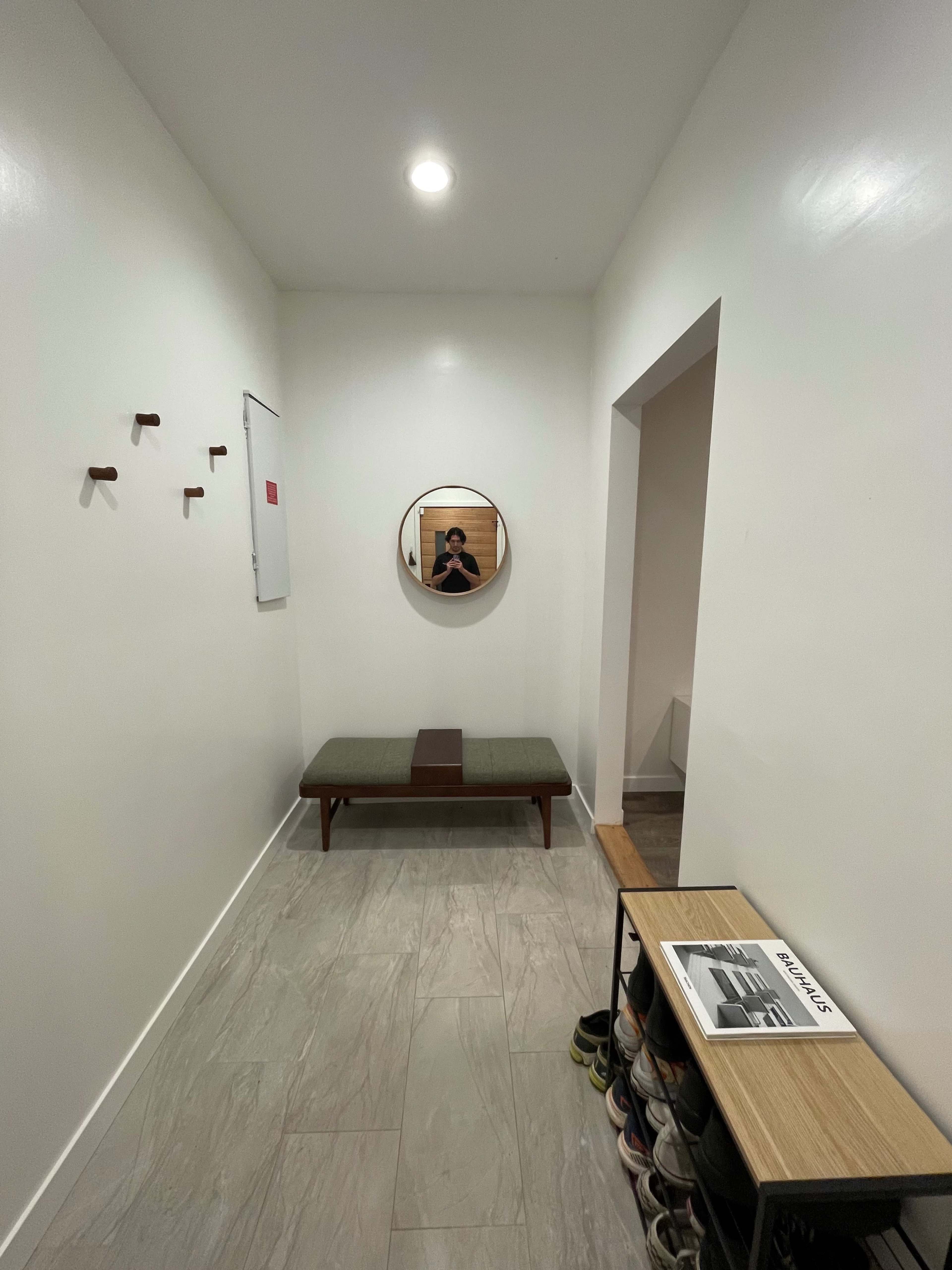 The image shows a narrow hallway with a round mirror, a bench, and a shoe rack against the wall.