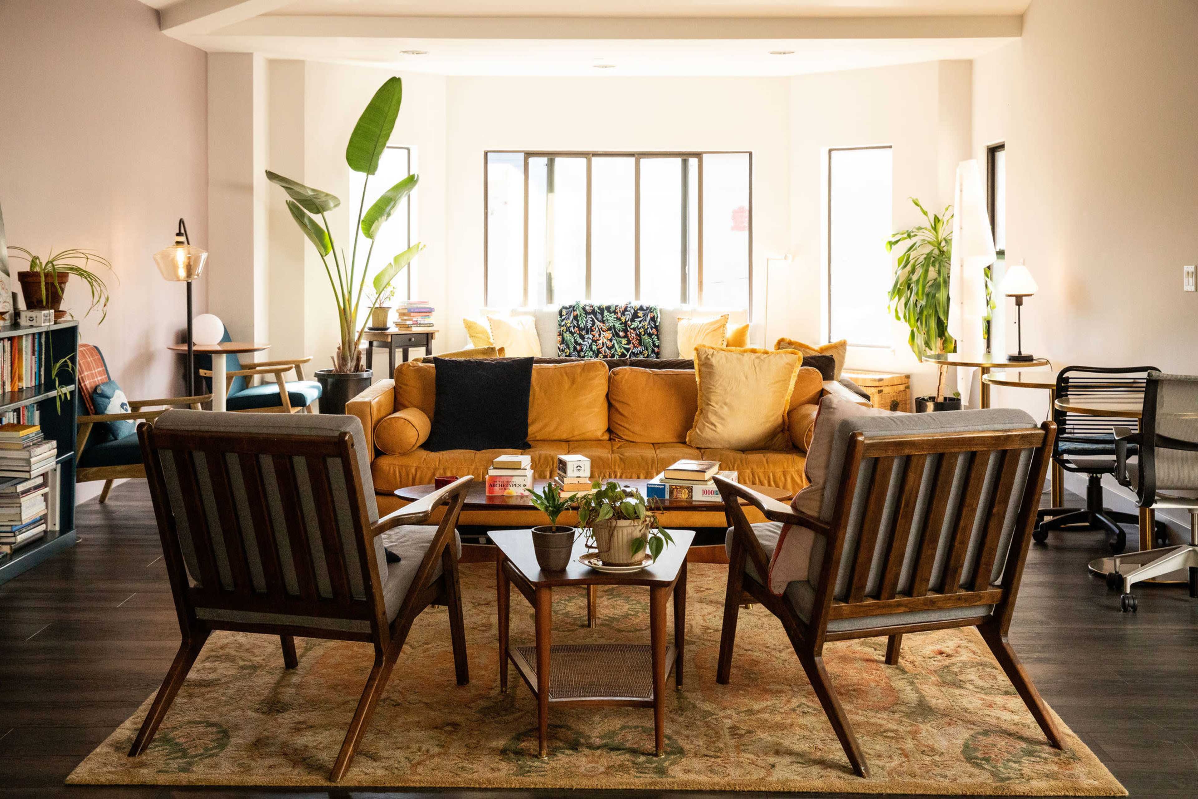 The image shows a modern living room with a yellow sofa, two wooden armchairs facing a coffee table, and large windows allowing natural light to enter the space.