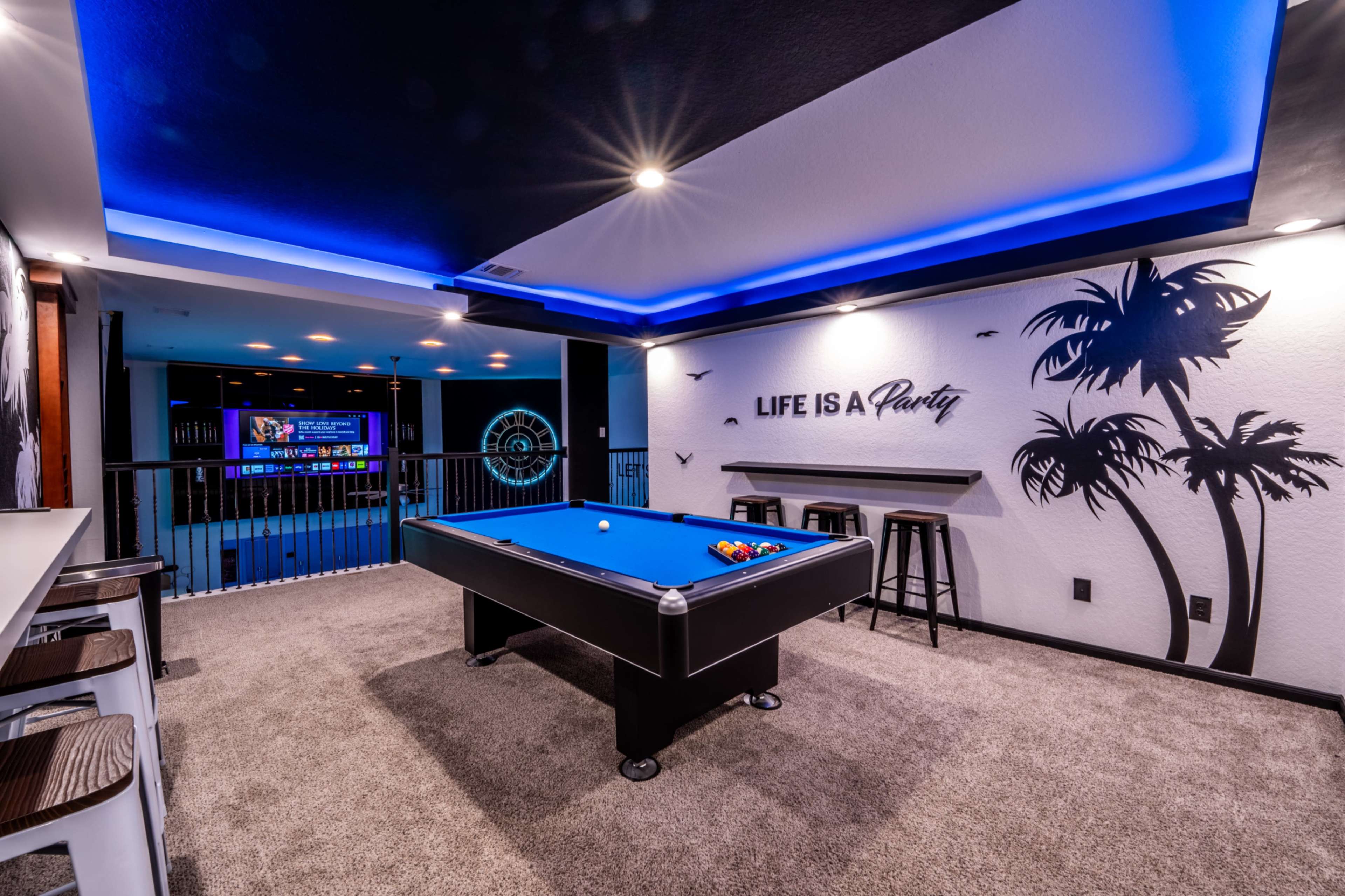 The image shows a modern game room featuring a blue felt pool table, bar stools, and a wall with a tropical theme and the phrase "LIFE IS A Party."