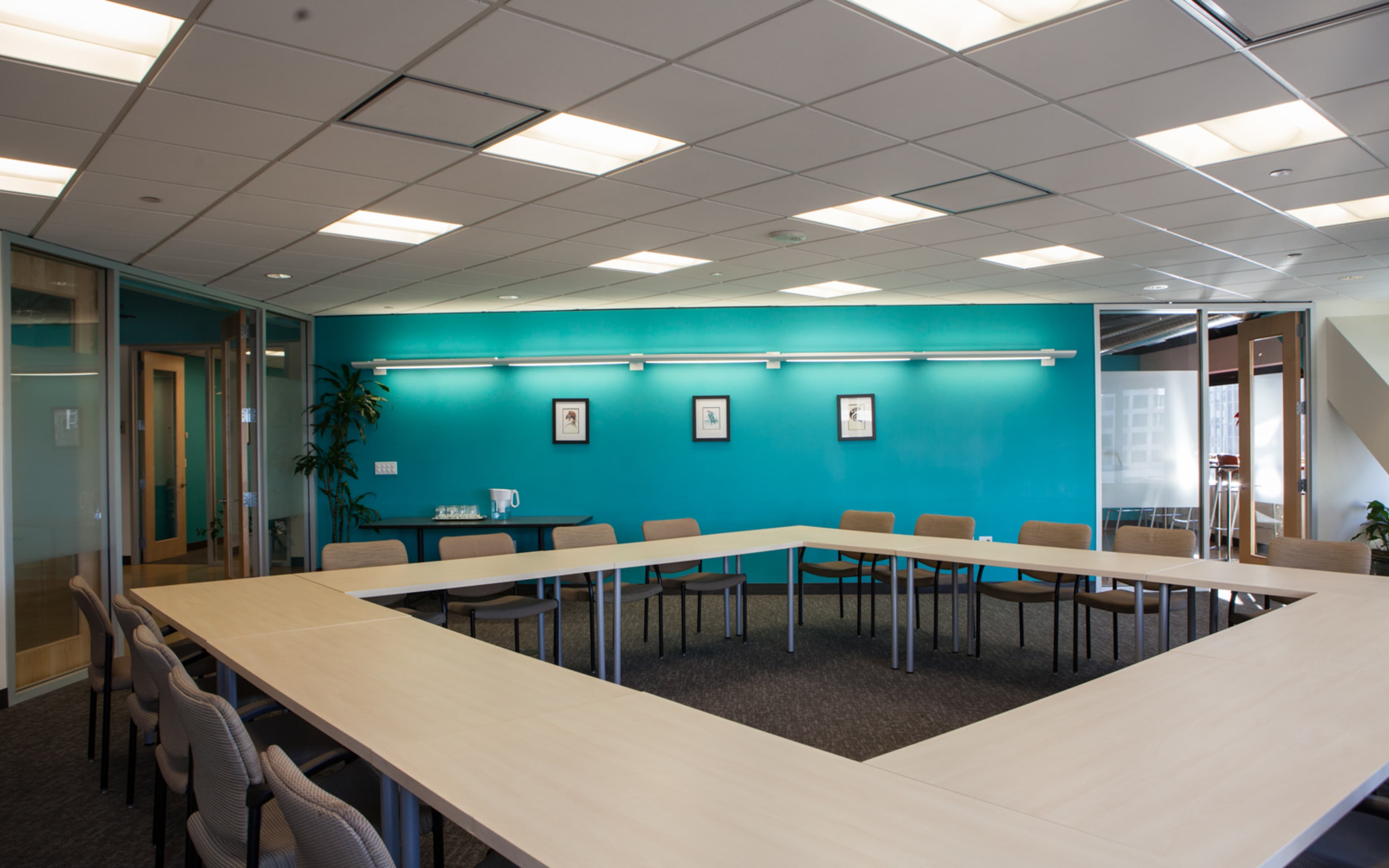 A conference room featuring a large, U-shaped table surrounded by chairs, with blue walls and a glass-walled meeting area.
