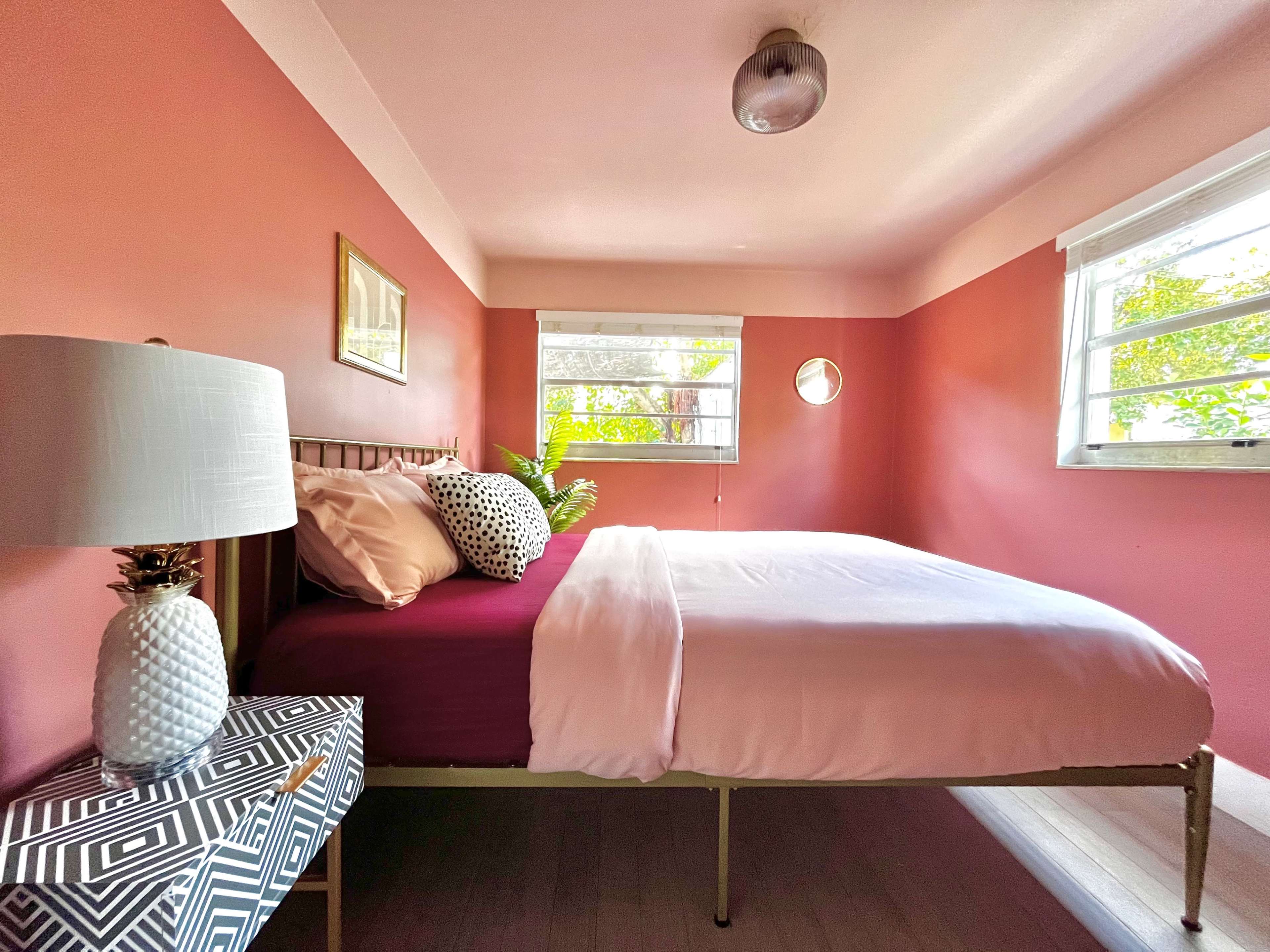 A bedroom with a maroon accent wall, a bed with white and patterned bedding, and a nightstand with a decorative lamp and plant.