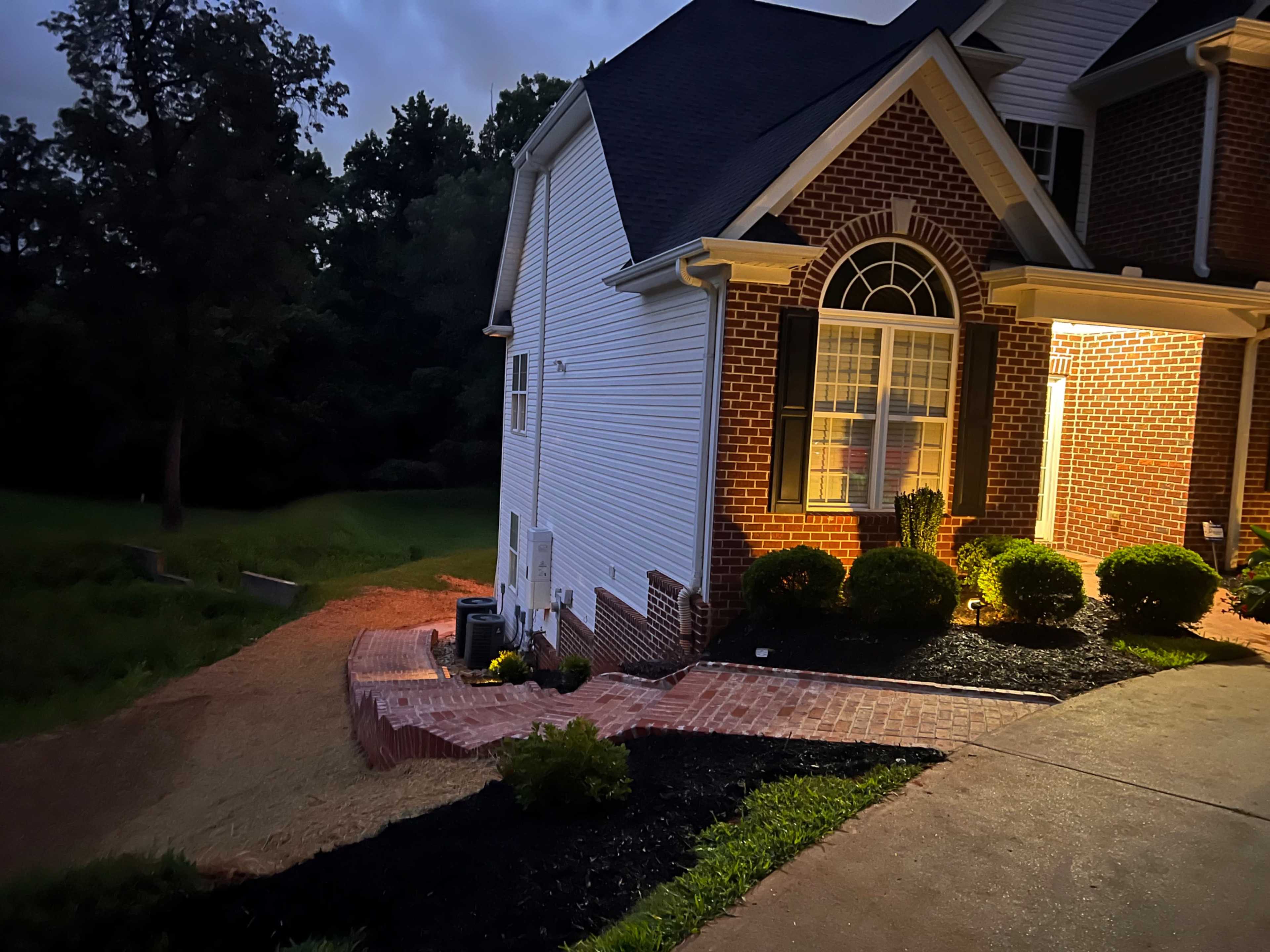 A brick walkway leading to a well-lit corner of a house with a landscaped yard at twilight.