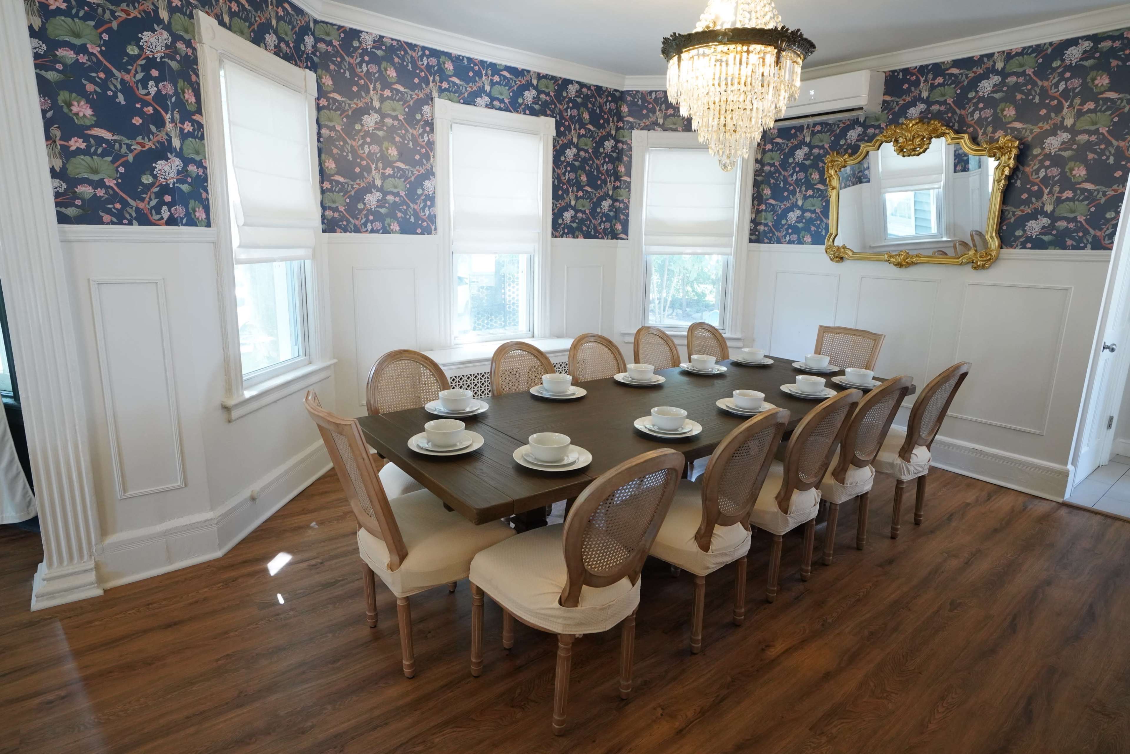 A long wooden dining table set for twelve is positioned in a room with floral wallpaper and a chandelier.