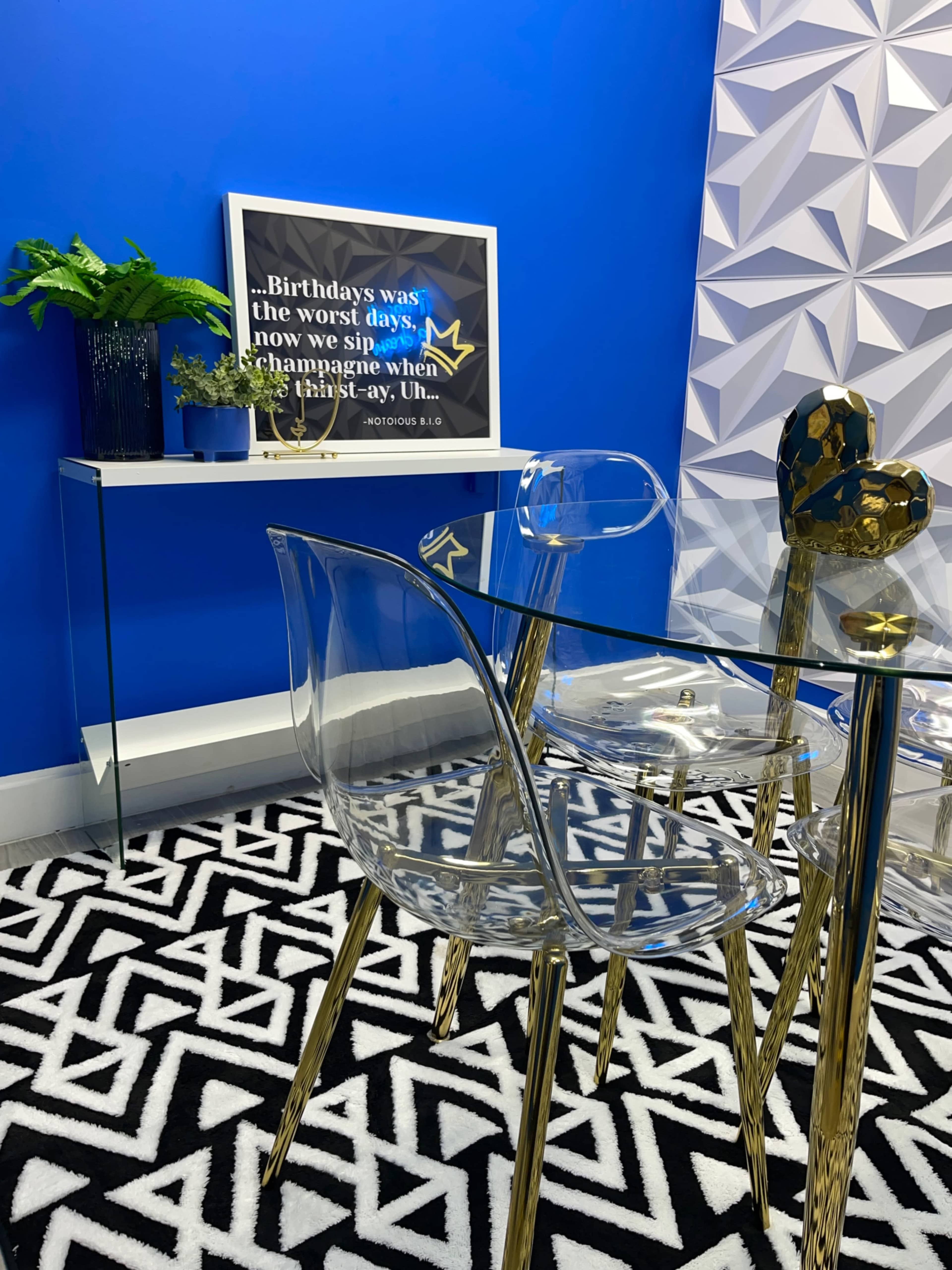 A modern dining area with a glass table and transparent chairs, a decorative wall art piece, and a patterned black and white rug on a blue background.
