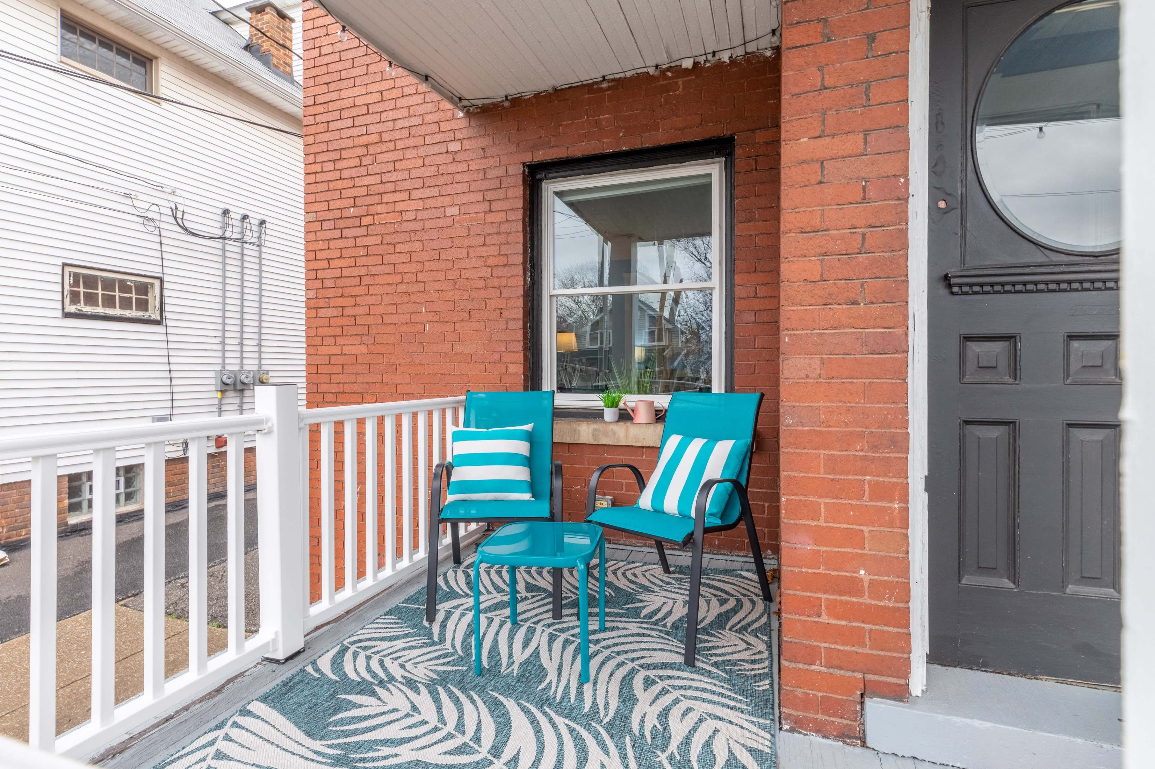 A small porch features two teal chairs and a matching table on a patterned rug, with a window adjacent to a front door.