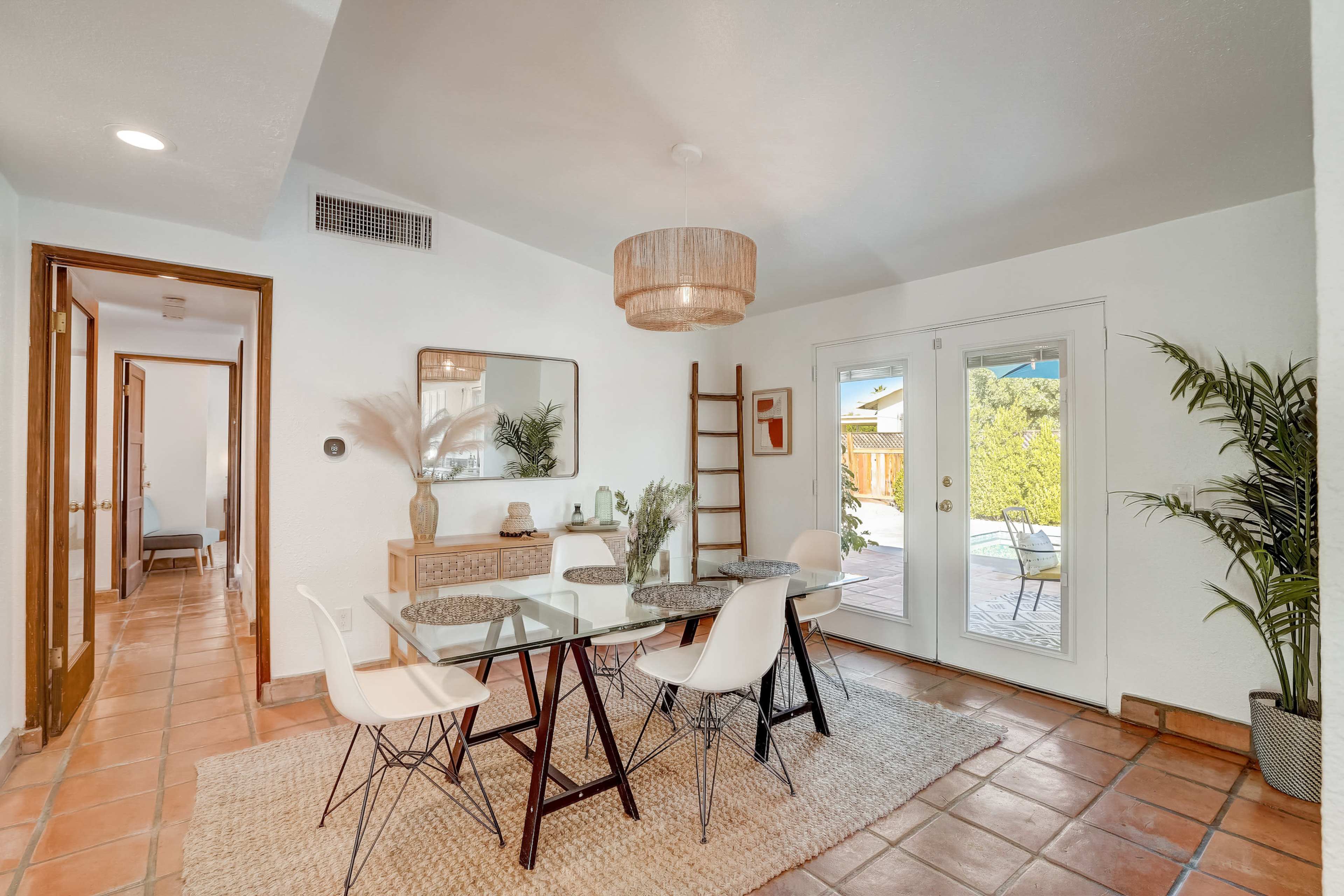 A dining area features a glass table with white chairs, a decorative mirror, and access to an outdoor patio through double doors.