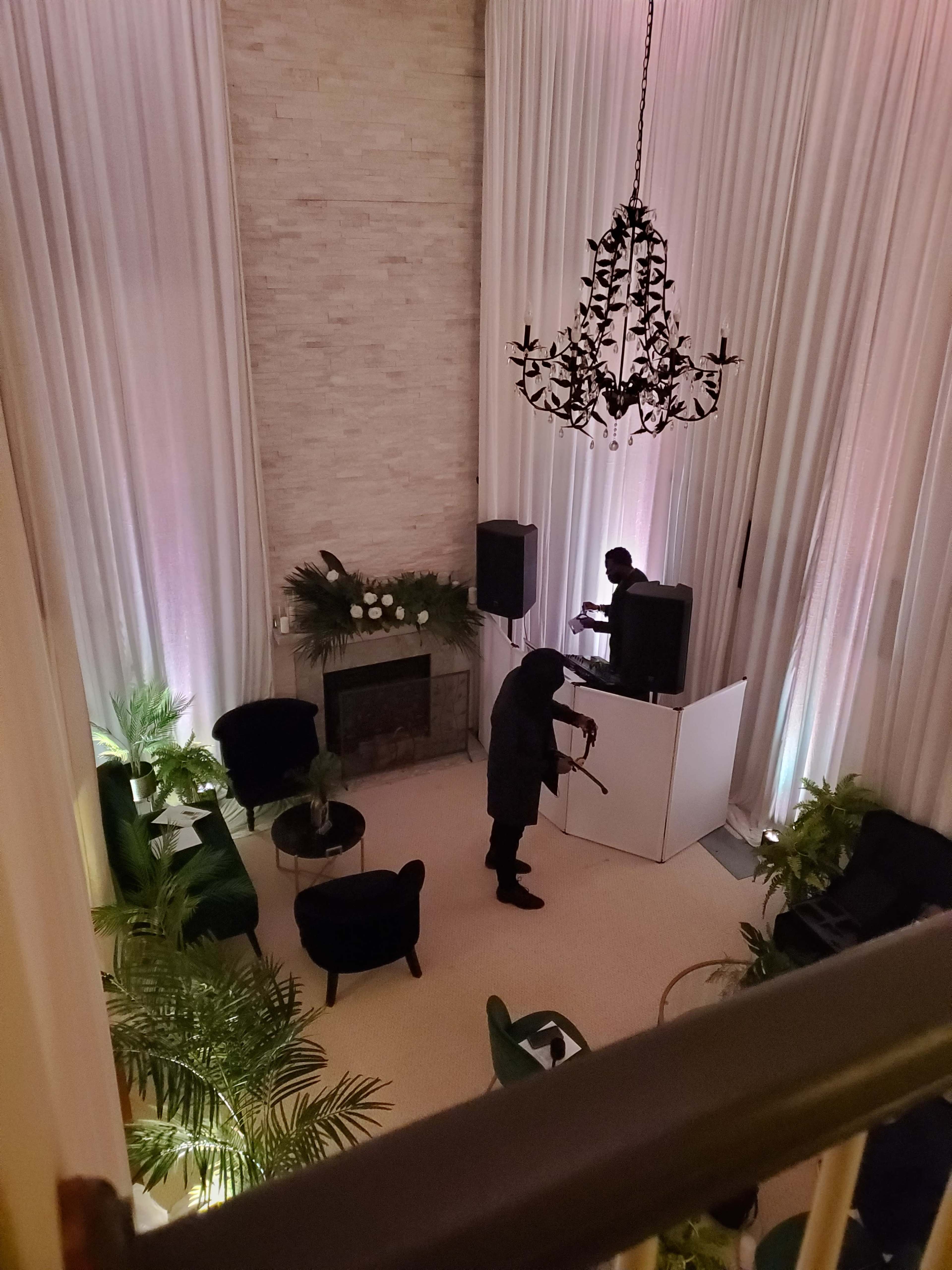 A person plays a violin in a spacious room with a fireplace, large curtains, and green plants, while another individual operates equipment at a DJ booth.